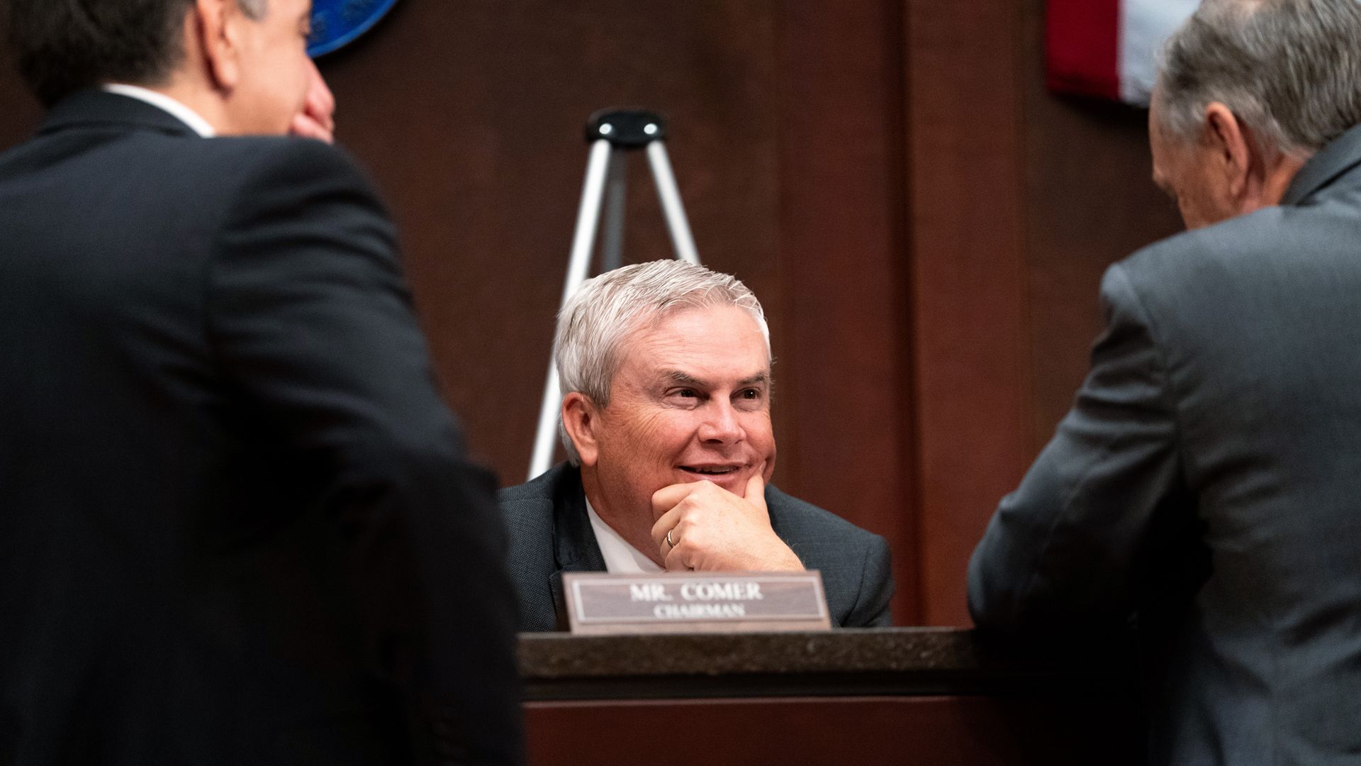 A man with gray hair labeled Mr. Comer, chairman, smiles as he rests his chin on his hand behind a desk, with two other men in suits in the foreground.