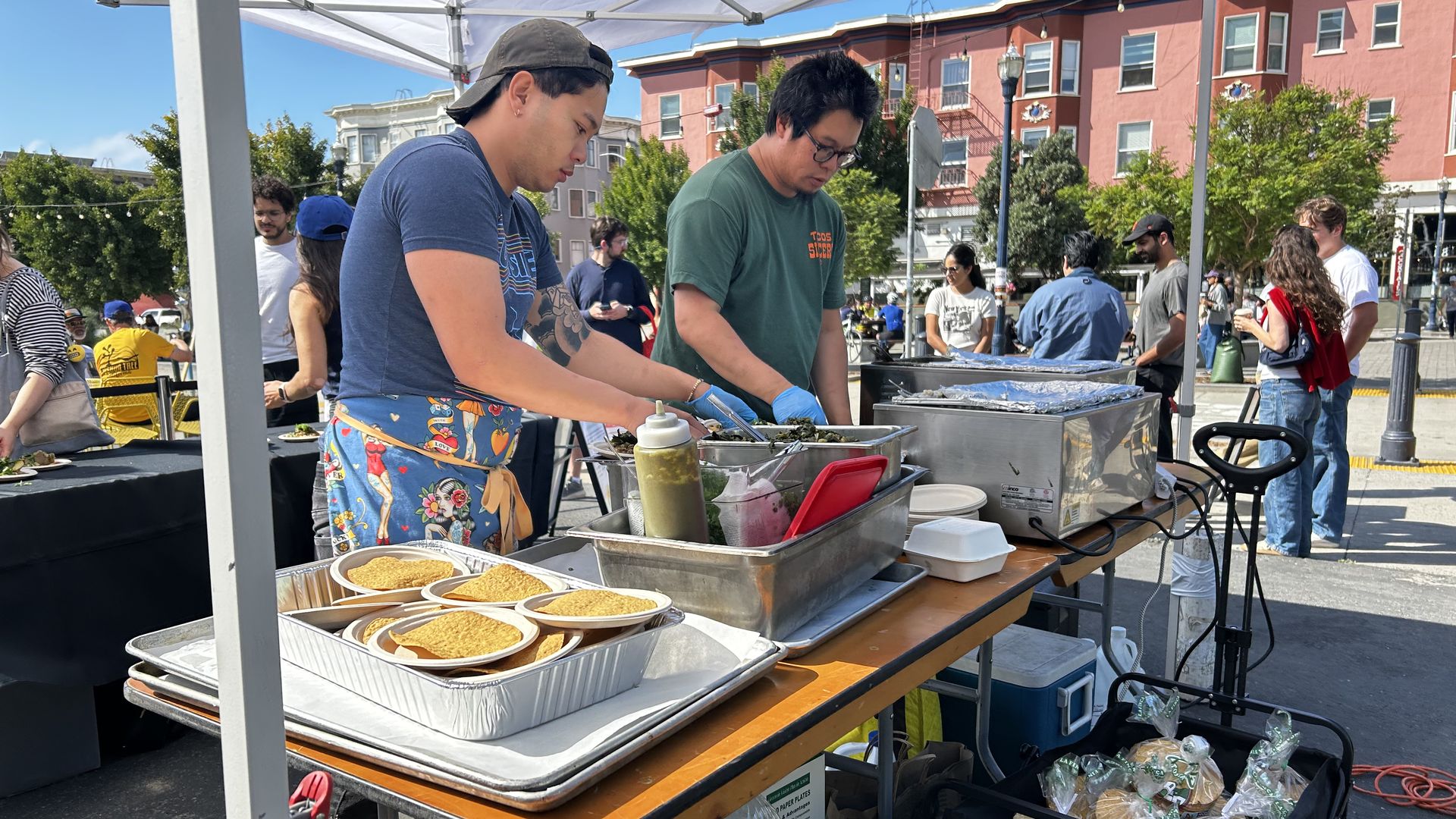 Two men preparing food at an outdoor market stand under a white tent, with trays of tacos and condiments on the table, and people socializing in the background near pink buildings.