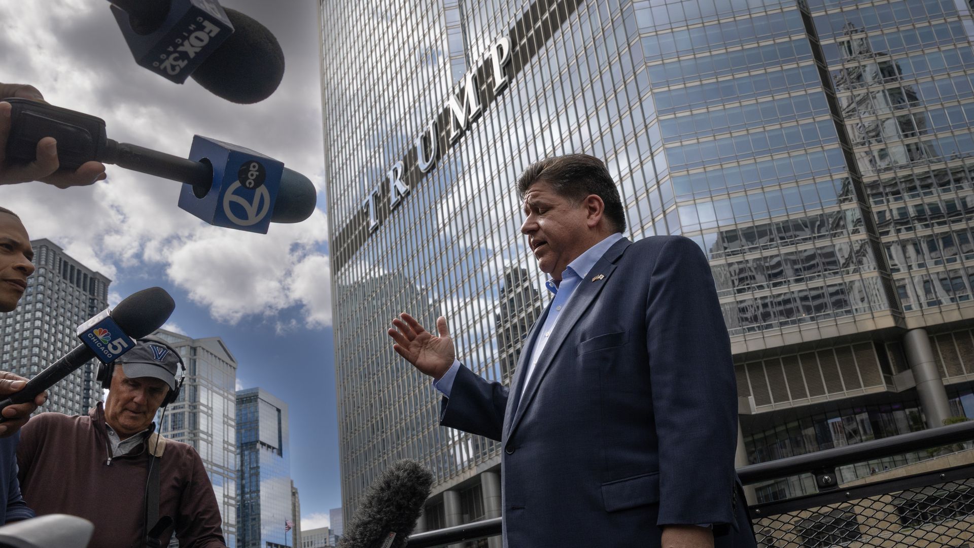 Illinois Gov. JB Pritzker, wearing a dark blue suit, speaks to reporters holding microphones labeled ABC 7, NBC 5 Chicago, and FOX 32 in front of the reflective Trump Tower building under a cloudy sky.