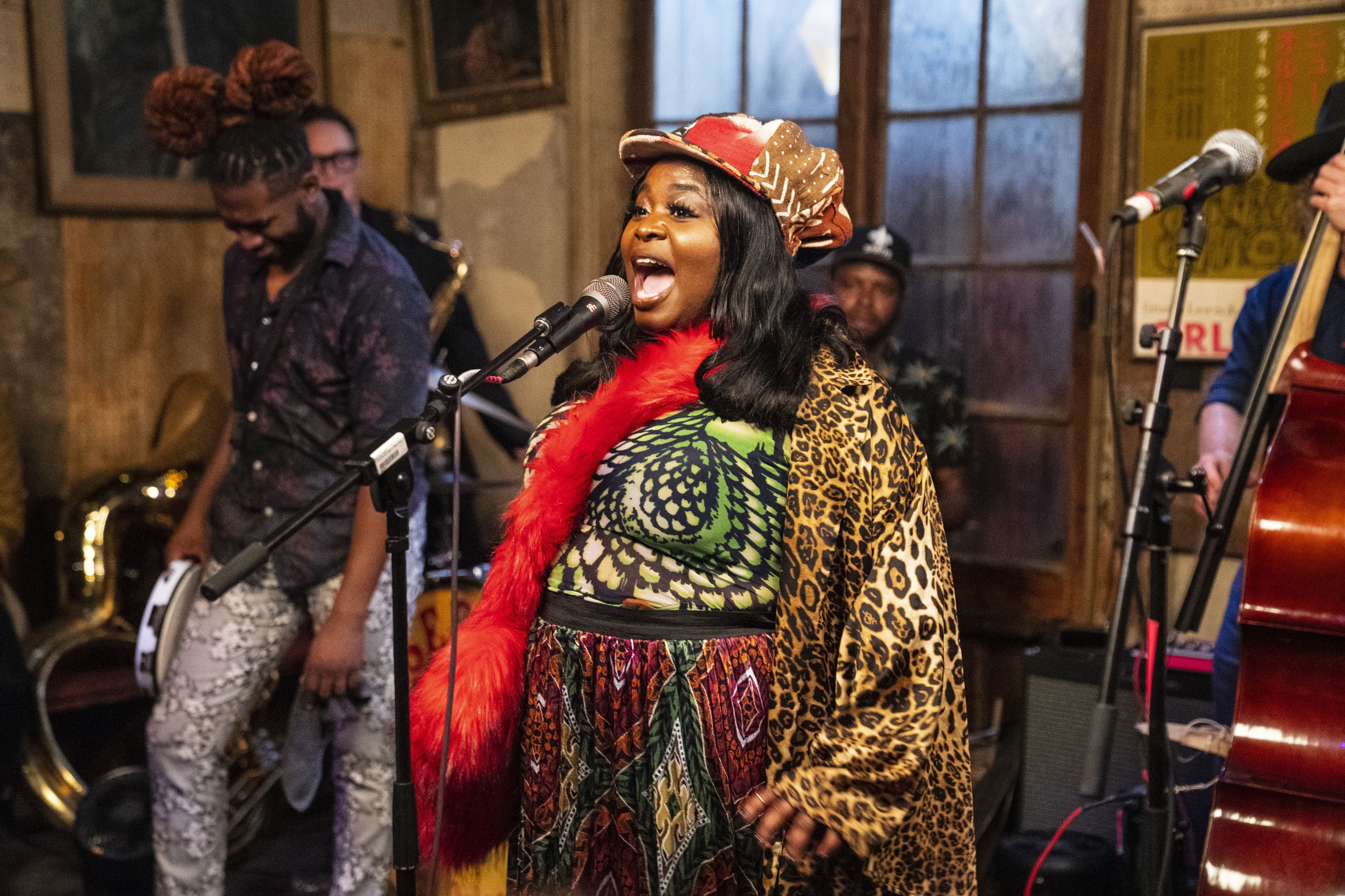 Tank Ball performs inside Preservation Hall. She stands at a microphone while other musicians can be seen behind her, including a bass player, drummer, and someone holding a tambourine.