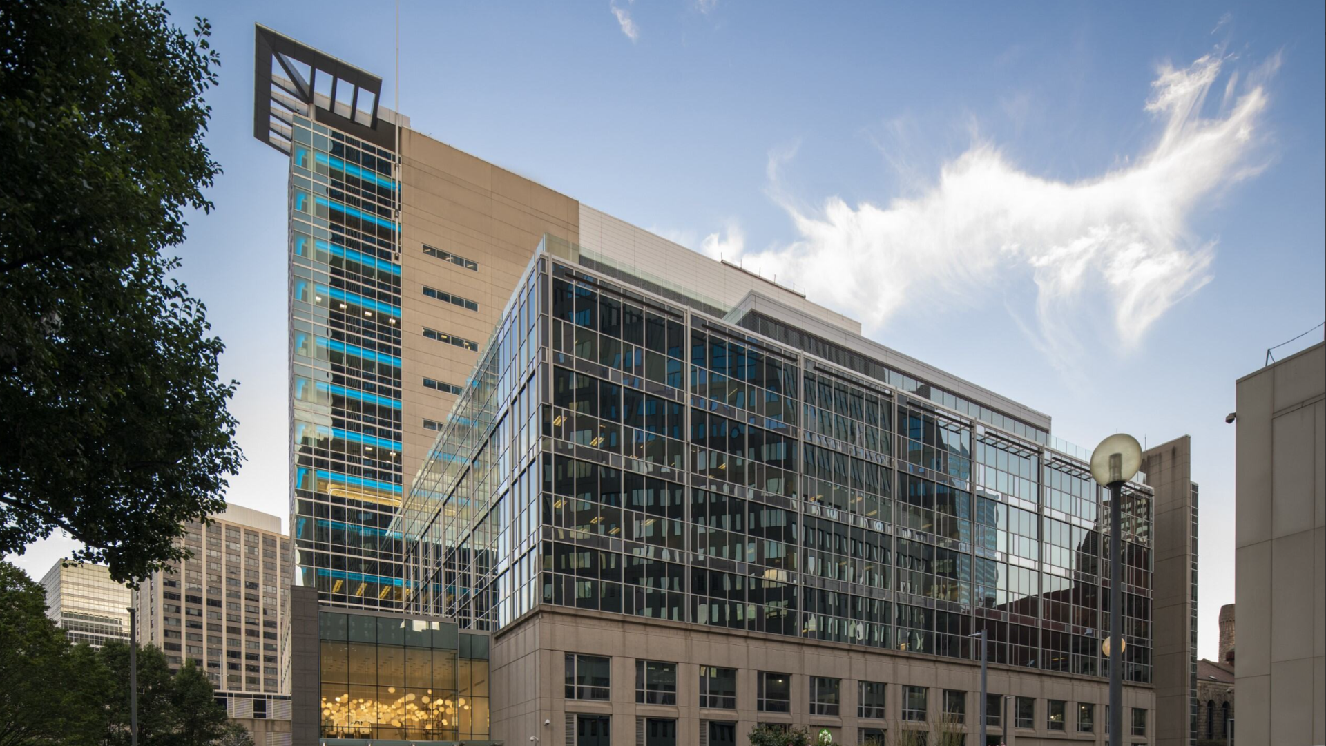 Modern office building with glass facade and beige concrete sections under a blue sky with a wispy white cloud, surrounded by other tall buildings and trees on a city street.