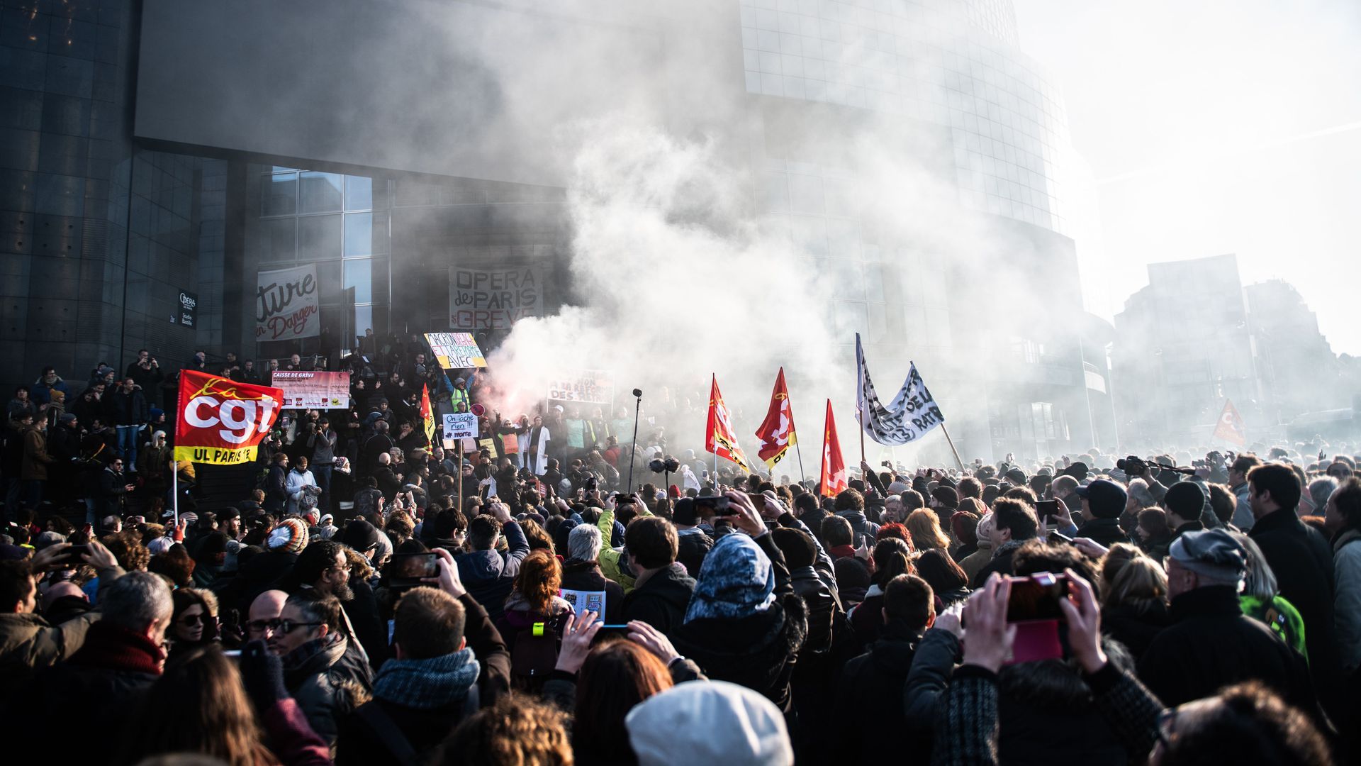 In this image, a large crowd of French protestors raise flags amid smoke