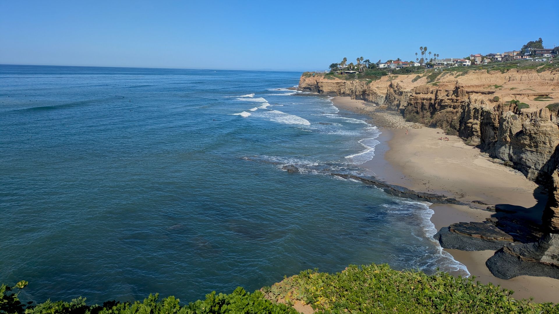 View of a sunny coastal cliff with beige rock formations, green shrubbery in the foreground, blue ocean waves, and a small sandy beach with houses and palm trees on top of the cliffs.