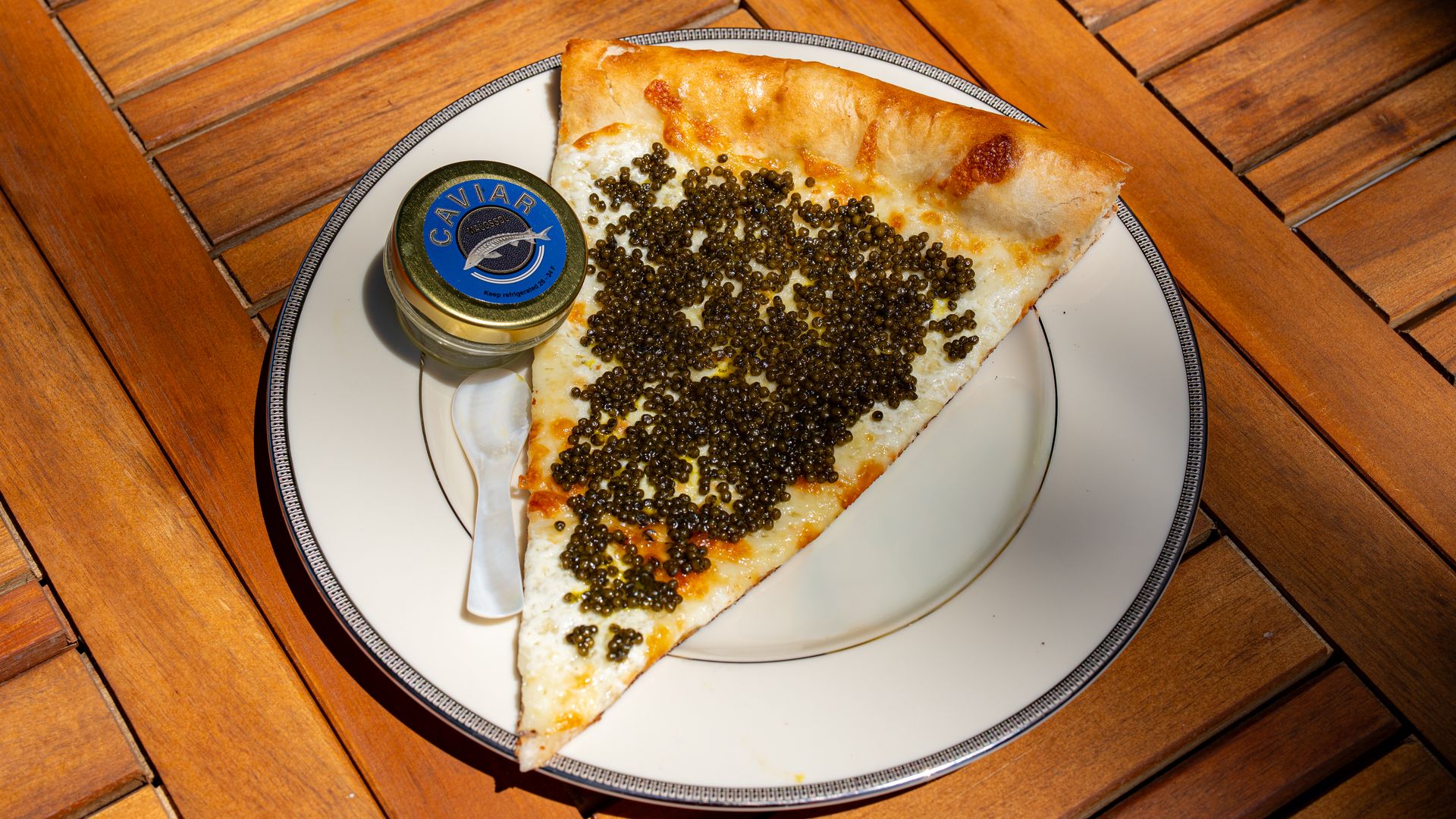 Slice of cheese pizza covered in black caviar on a white plate with a small jar labeled CAVIAR and a spoon, set on a wooden table.