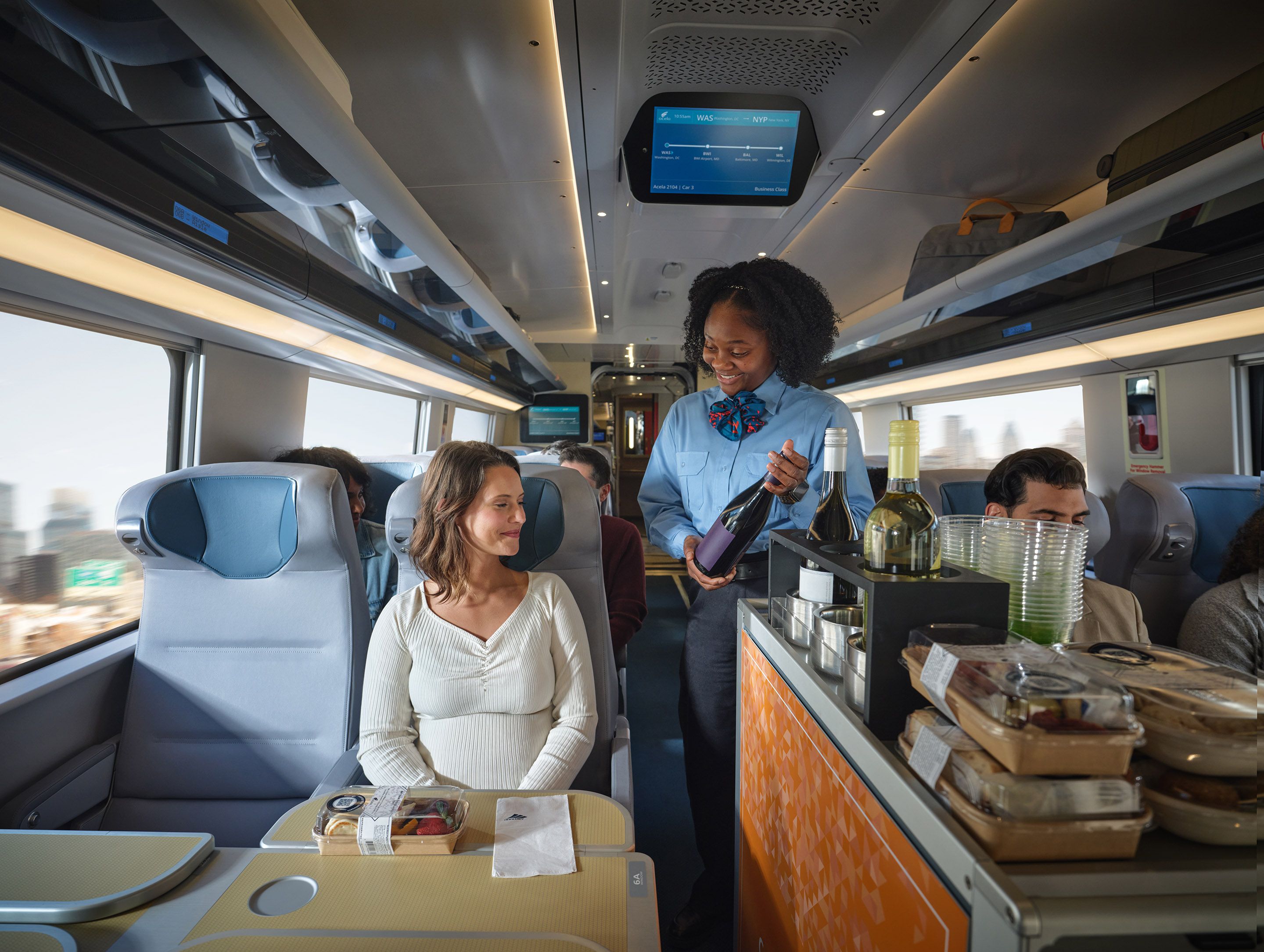 A smiling train attendant in a blue uniform serves a passenger in business class, holding a bottle of wine. The passenger, a woman in a white top, sits at a table with snacks on a modern train.
