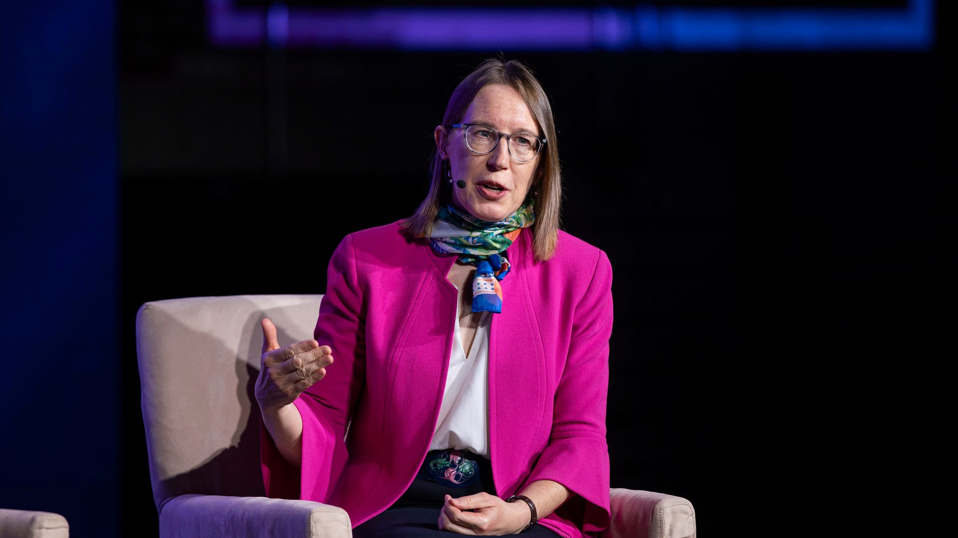 Woman in a pink blazer and scarf sitting on a conference chair speaking, gesturing.