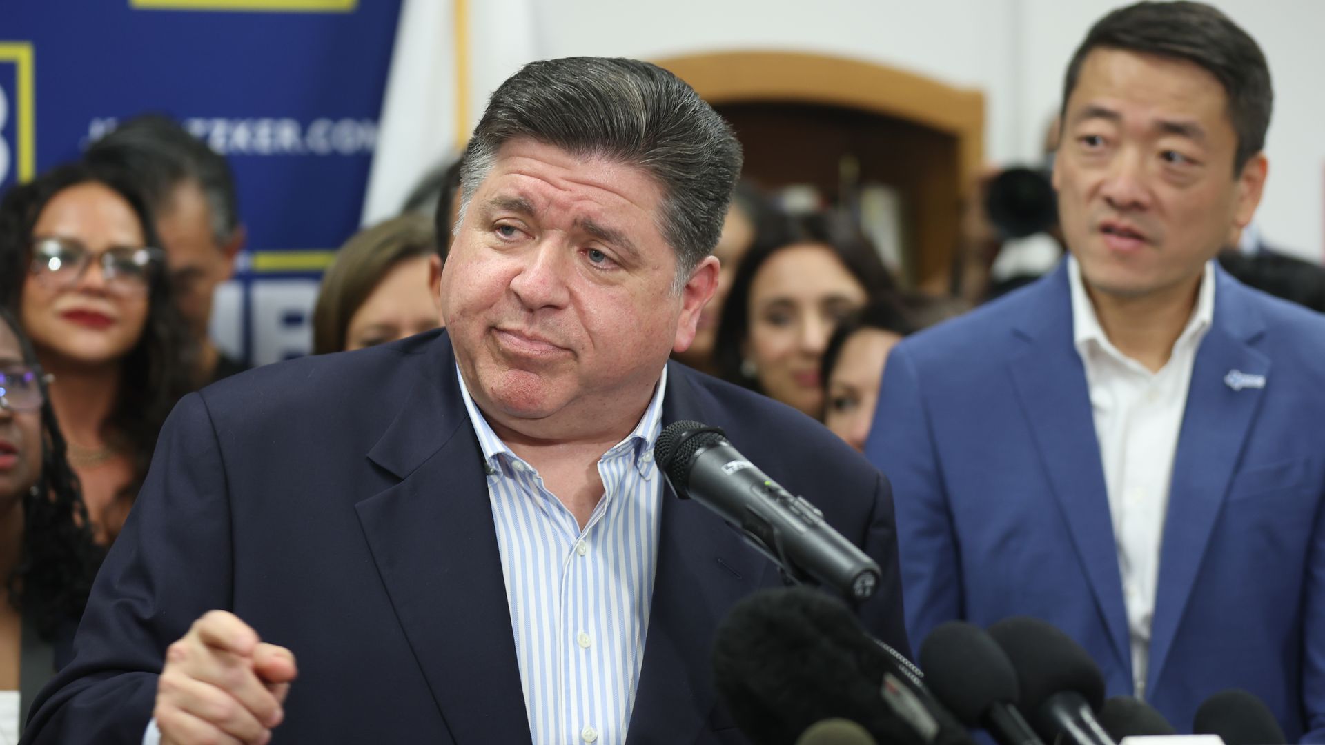A man in a dark suit with a striped shirt speaks into microphones at a press event, with people standing closely behind him and a man in a blue suit looking on to the right.