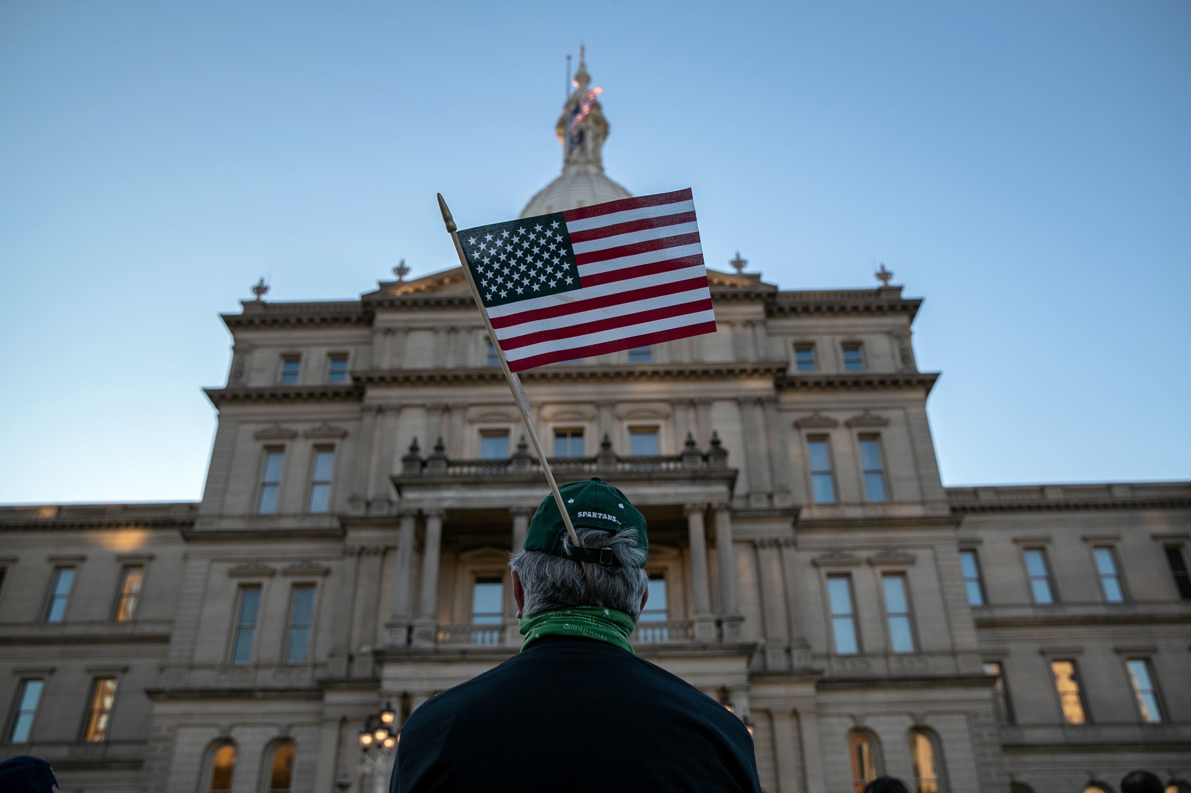 Person wearing a green hat with an American flag attached, standing in front of a large stone government building with many windows and a clear blue sky.