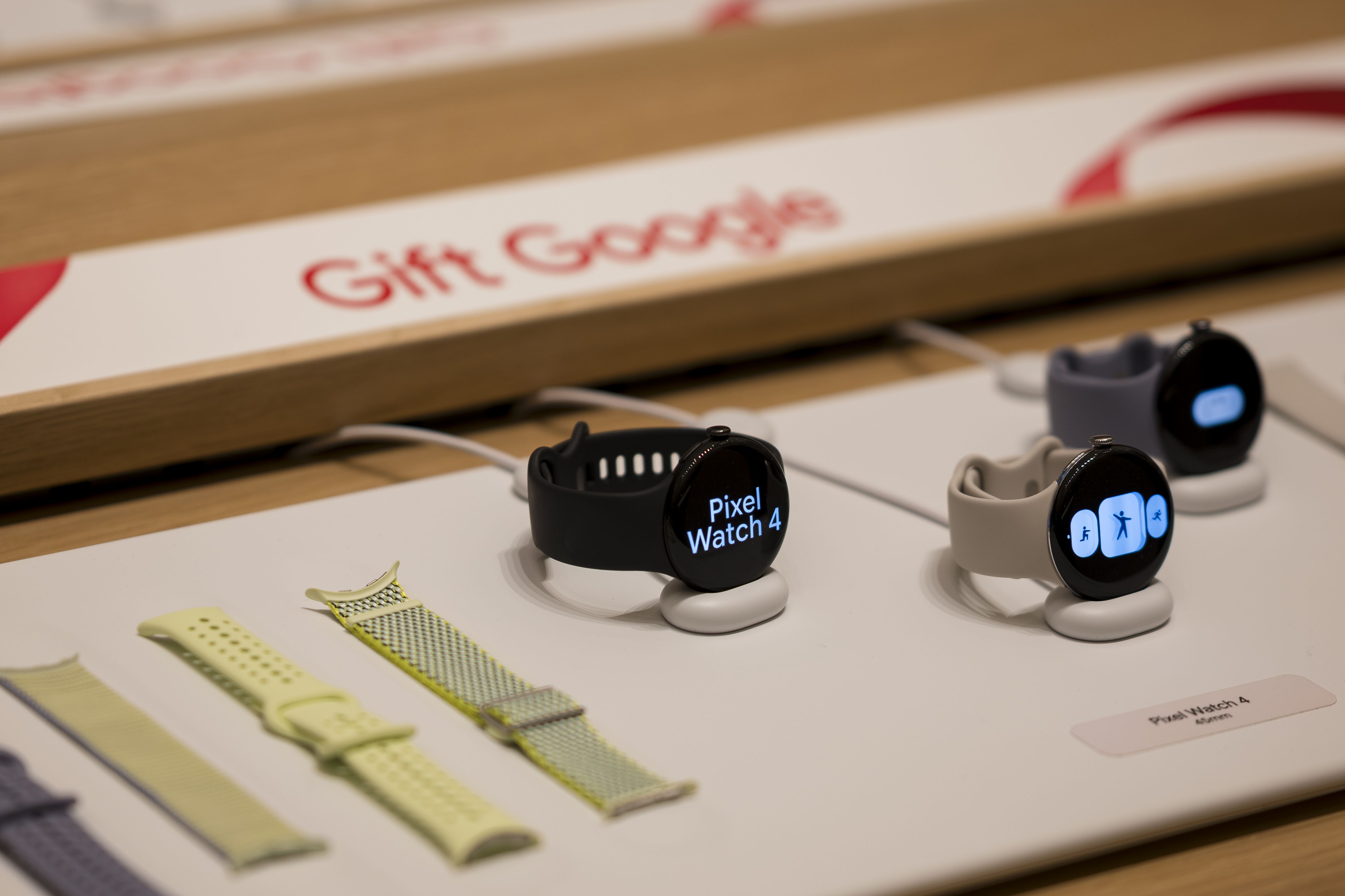 Display of Google Pixel Watch 4 smartwatches in black, beige, and blue with interchangeable bands in light green and yellow on a white showcase with "Gift Goods" sign in background.