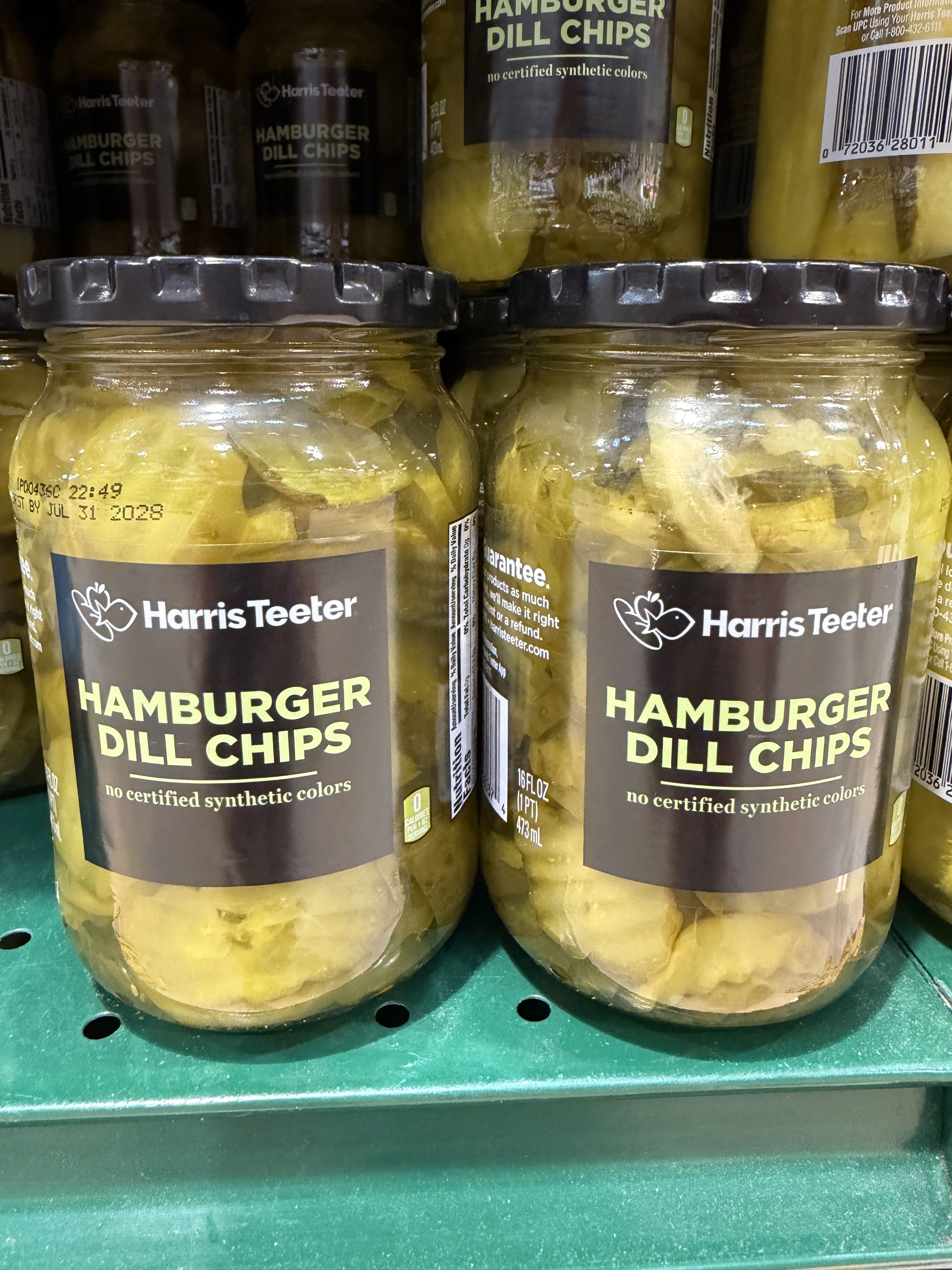 Two Harris Teeter hamburger dill chips jars on a grocery shelf, filled with yellow pickles and black lids; label reads "Hamburger Dill Chips".