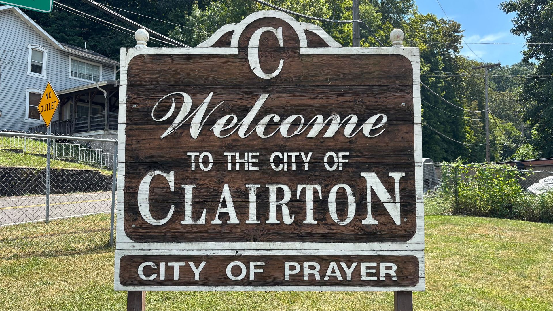 Wooden welcome sign for Clairton city with white text reading "Welcome to the city of Clairton, City of Prayer," set against houses, chain link fence, and trees on a sunny day.