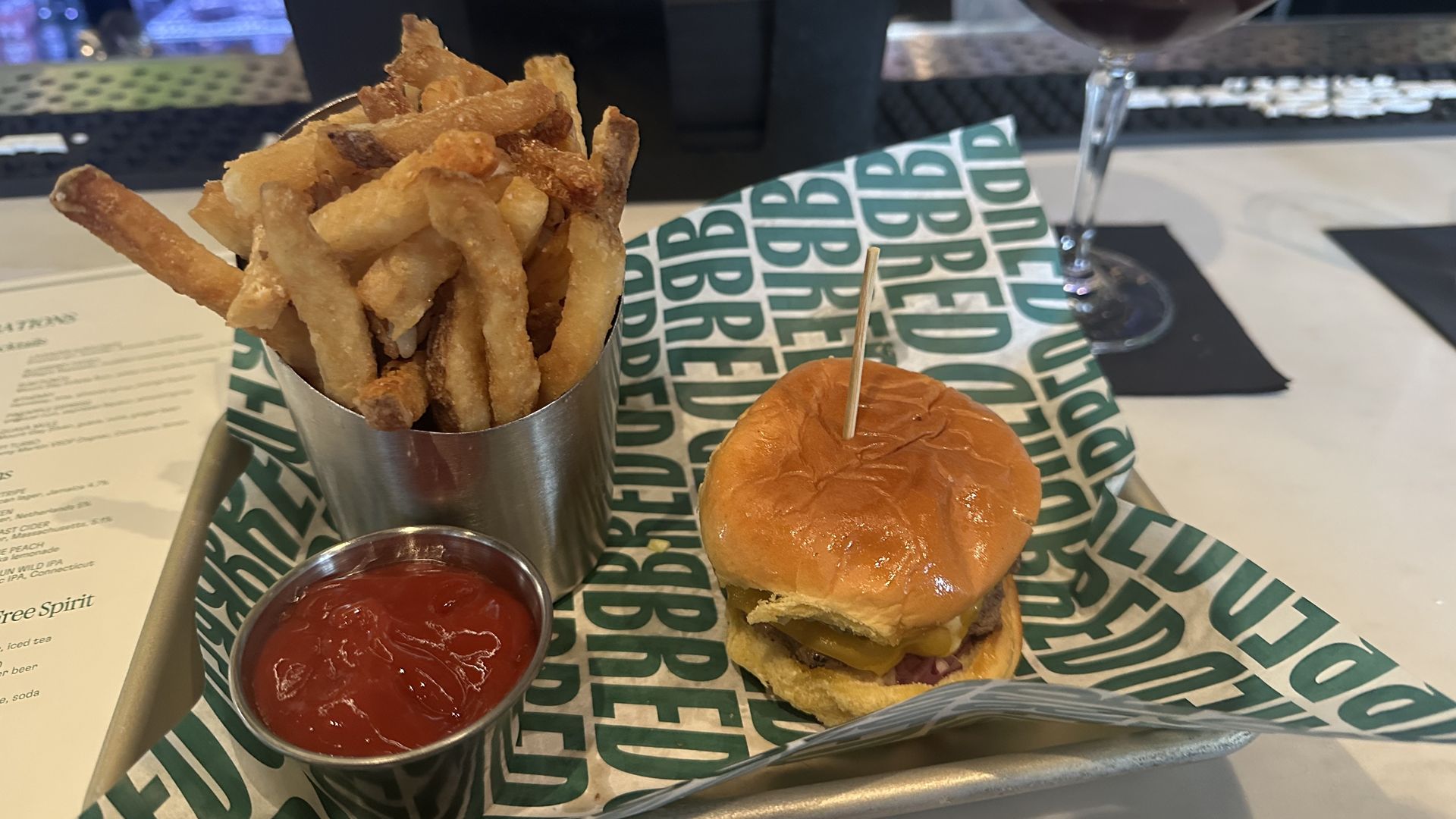 Tray with a cheeseburger slider on green and white paper, metal cup of fries, small cup of ketchup, and a dark cocktail with frothy top in a coupe glass on a bar counter.