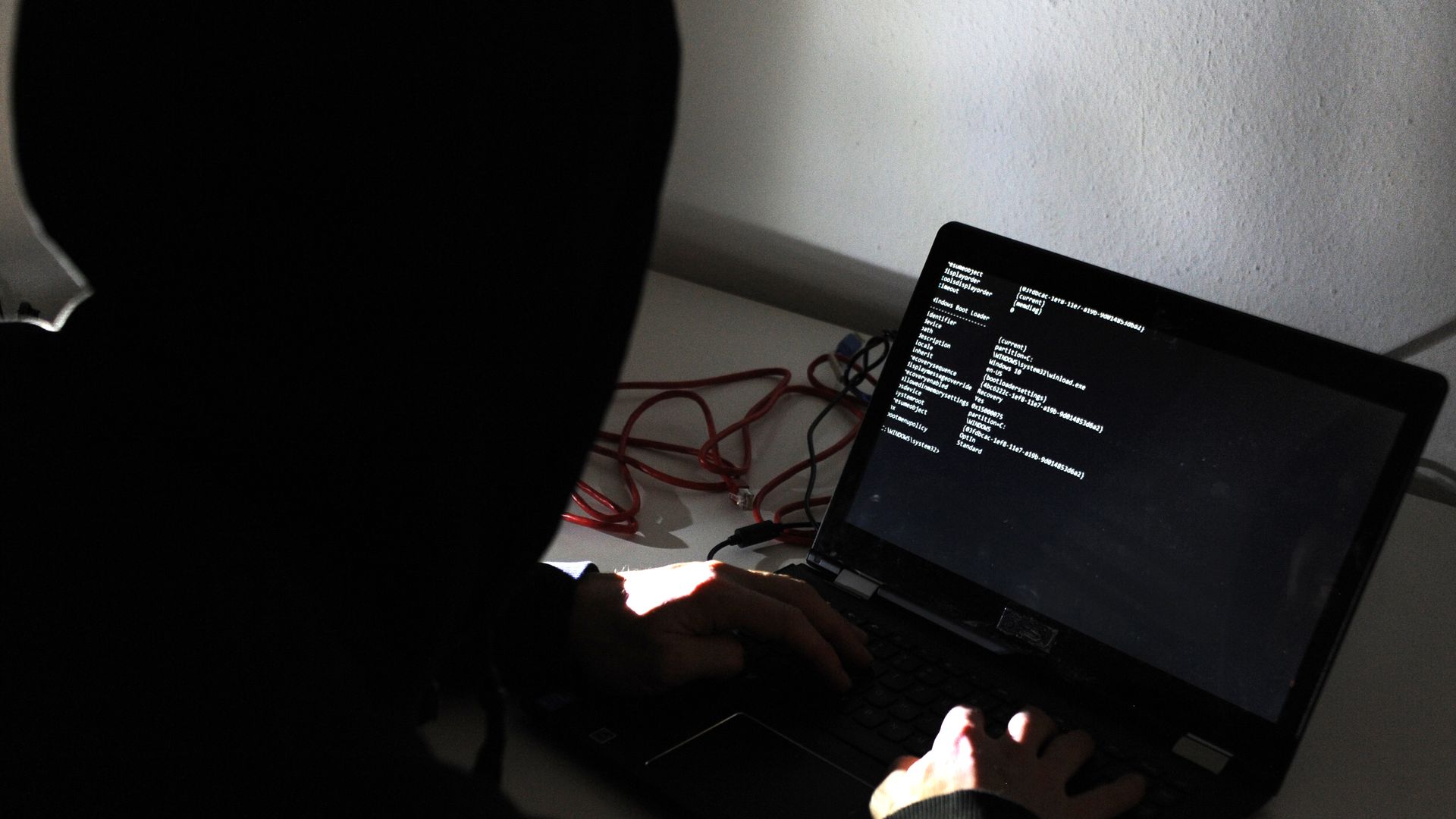 A man in a hoodie types on a black computer in darkness.