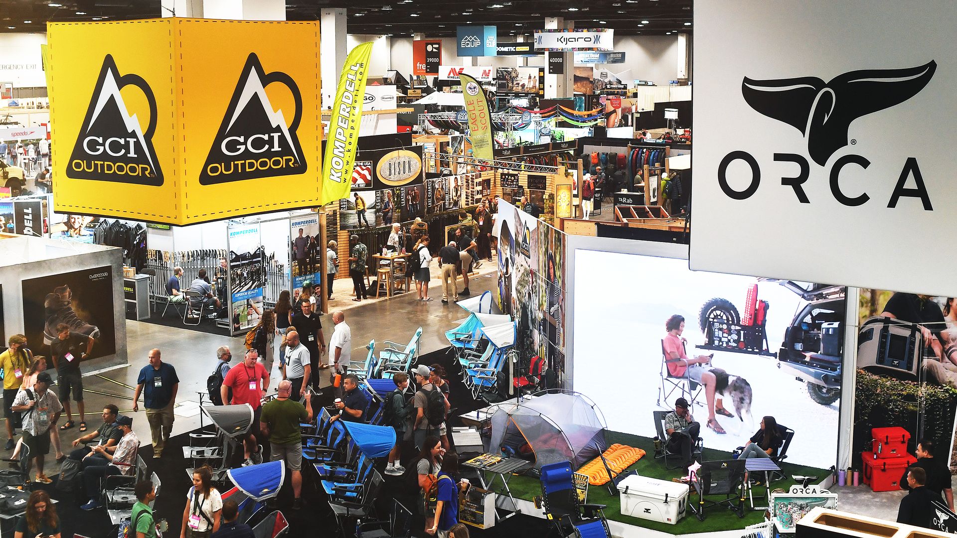 A crowded indoor outdoor gear expo with bright yellow GCI Outdoor banners left, a large ORCA sign right, rows of camping chairs and tents, booths, and many attendees browsing.
