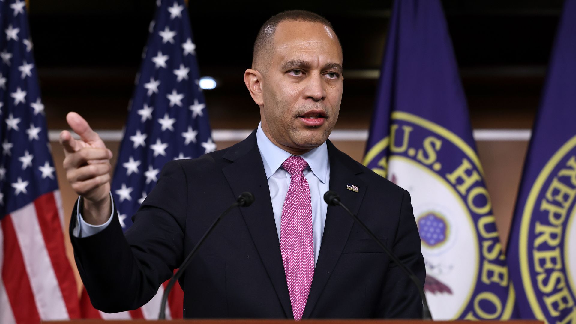 House Minority Leader Hakeem Jeffries wearing a blue suit and standing in front of a row of flags.