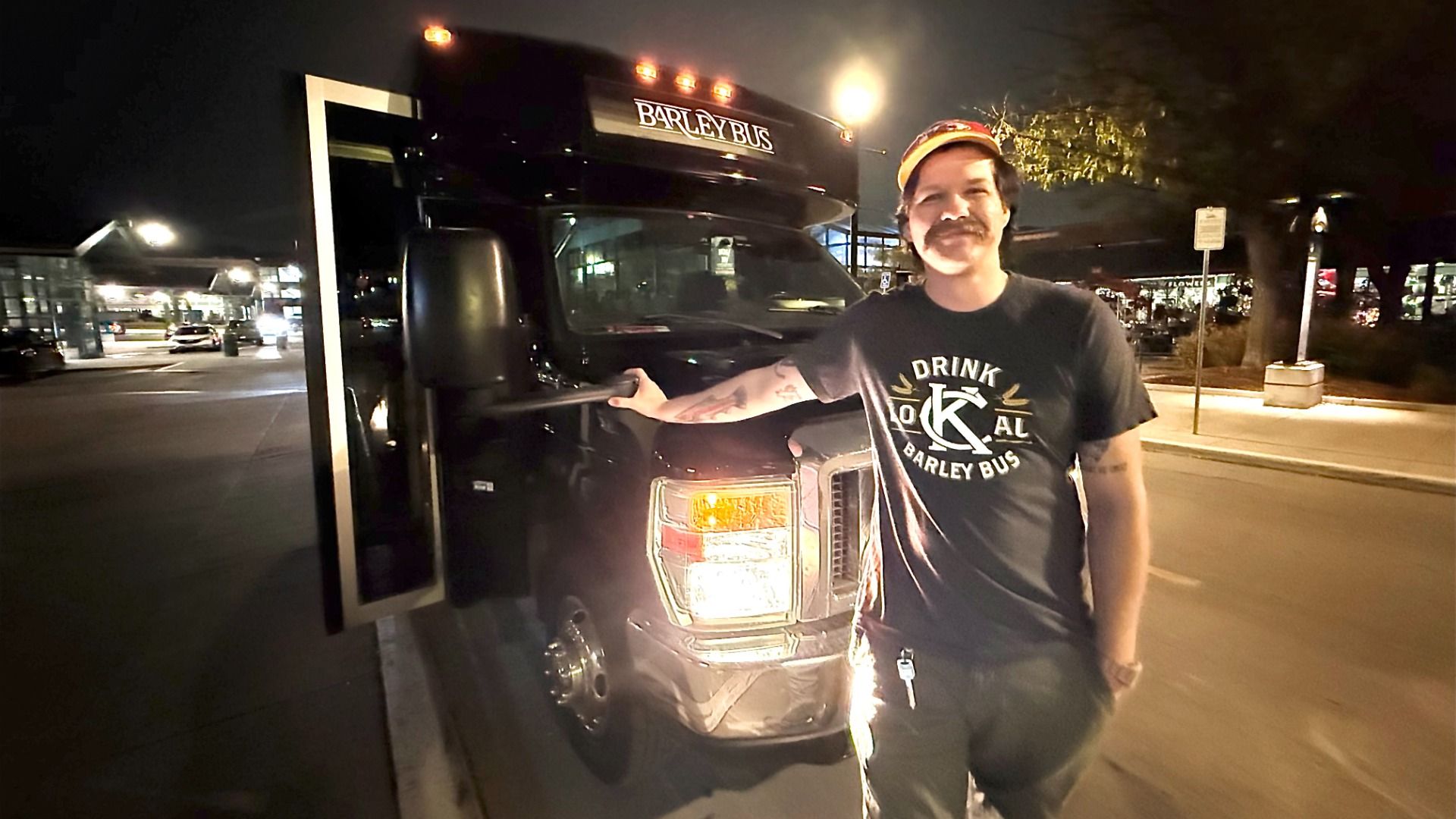 Man in black t-shirt and orange cap smiles, standing beside a black bus labeled "BARLEY BUS" at night, holding the bus door handle in a dimly lit urban street setting.