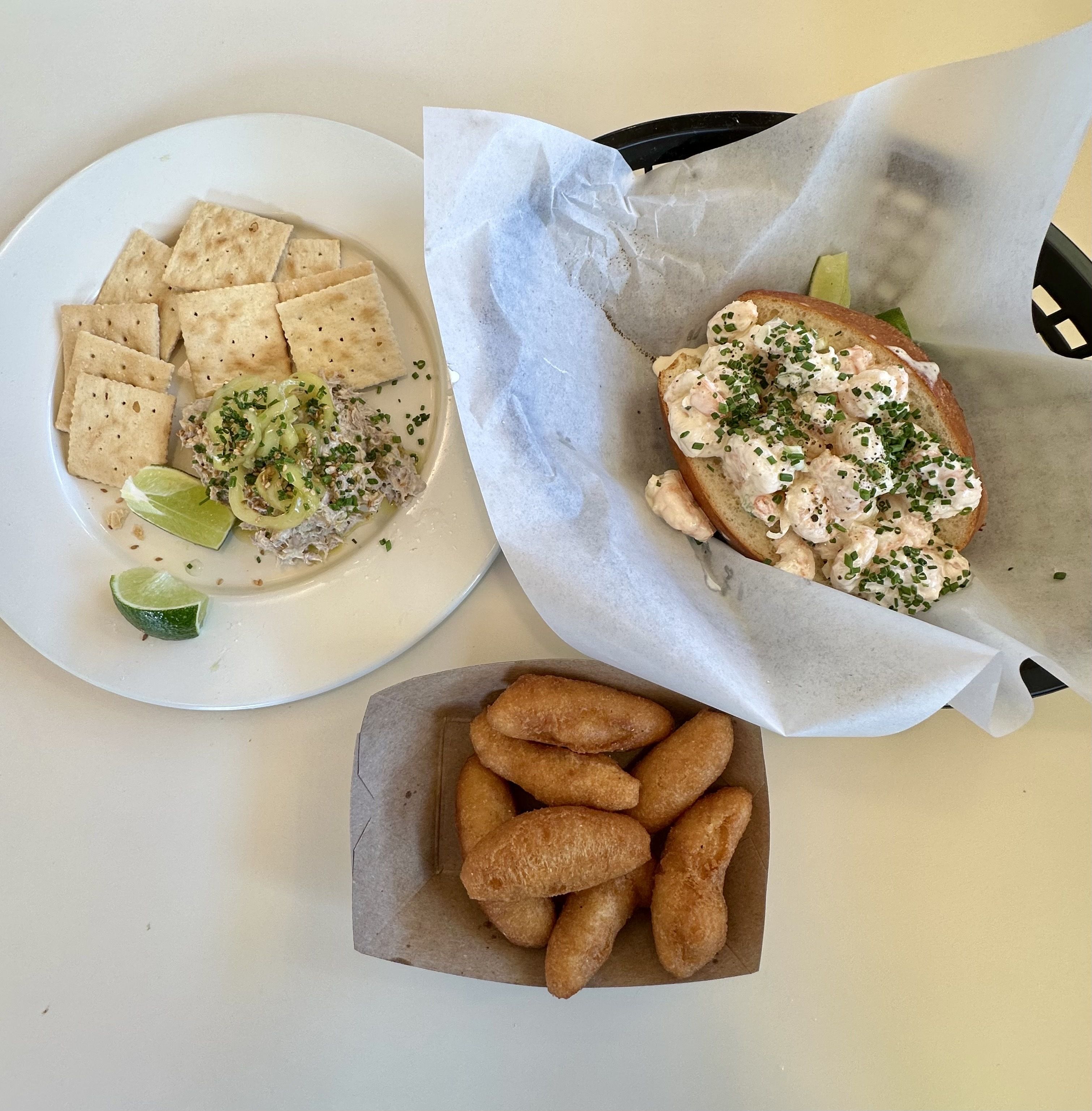 Top-down view of a meal: left plate with crackers and a lime-topped tuna/crab spread garnished with chives; right a lobster roll; small basket holds golden fried croquettes.