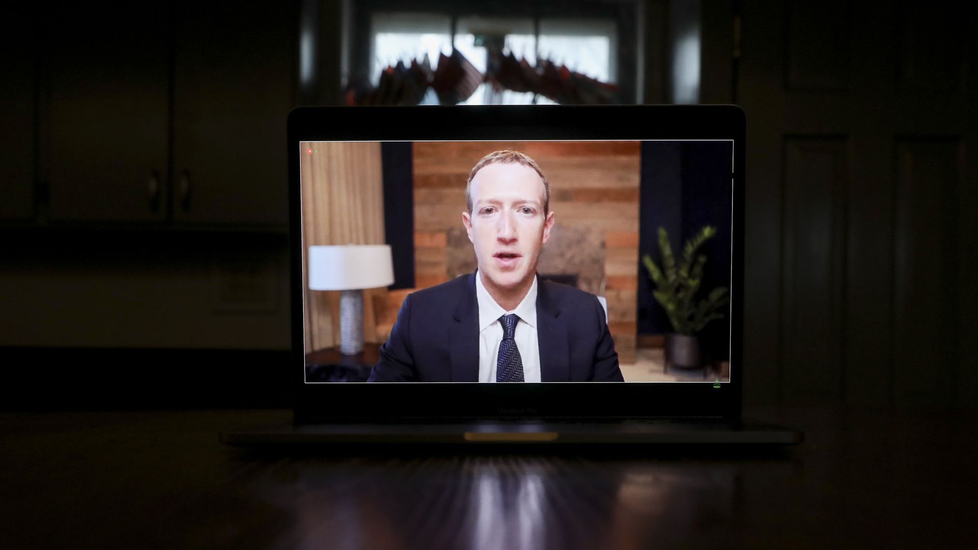 A man in a suit and tie speaking, seen on a tablet device placed on a table in a darkened room.
