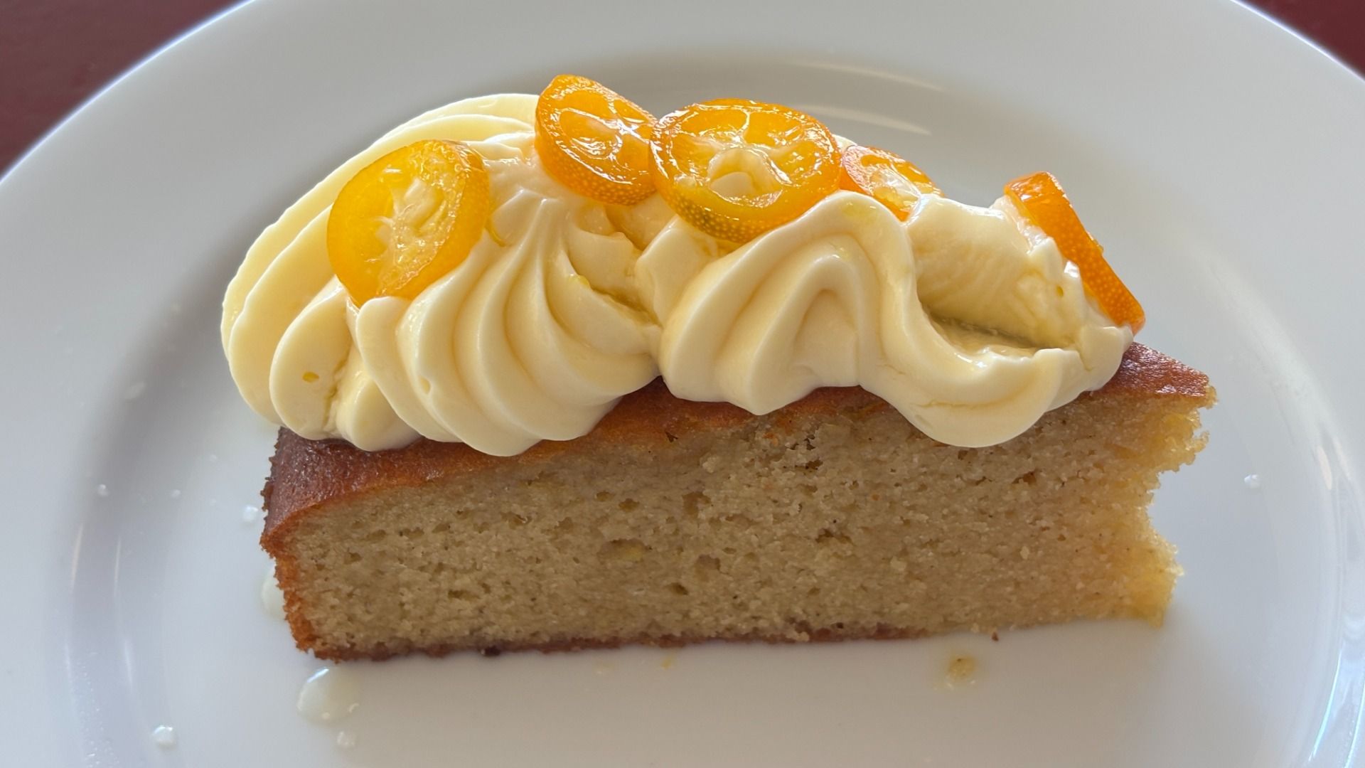 Slice of golden yellow cake with thick white buttercream frosting swirls topped with small orange slices, on a white plate.