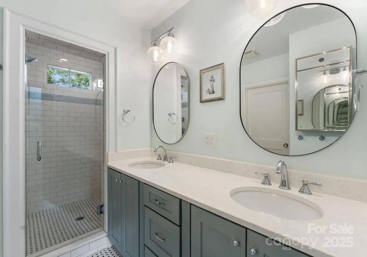 Modern bathroom with double sinks, green cabinets, oval mirrors, and glass-enclosed shower with white subway tiles and a small window.