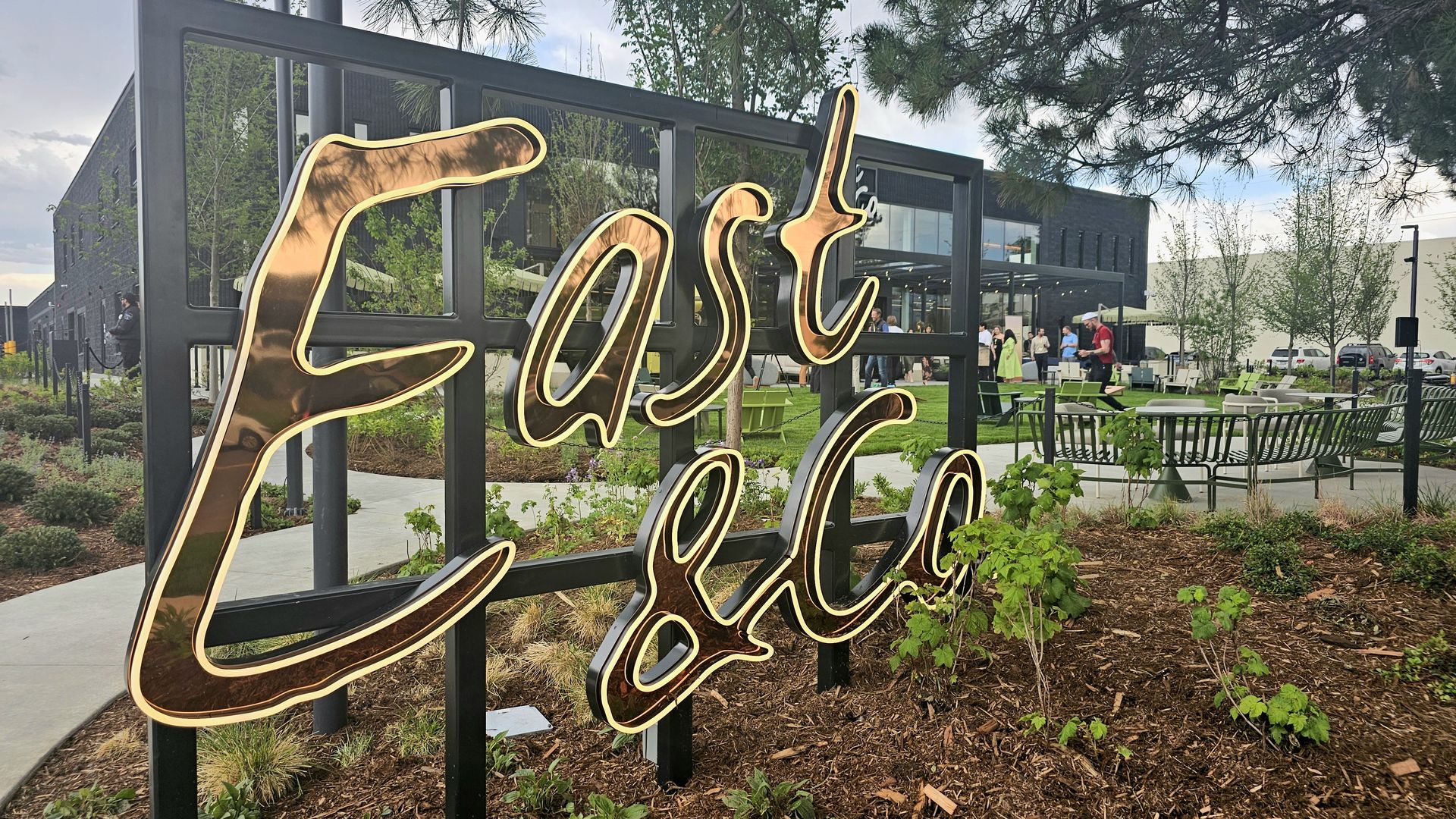 Outdoor scene featuring a large gold neon sign spelling Eat on a black frame; in the background is a modern building, outdoor seating, people mingling, trees, and landscaped garden beds.