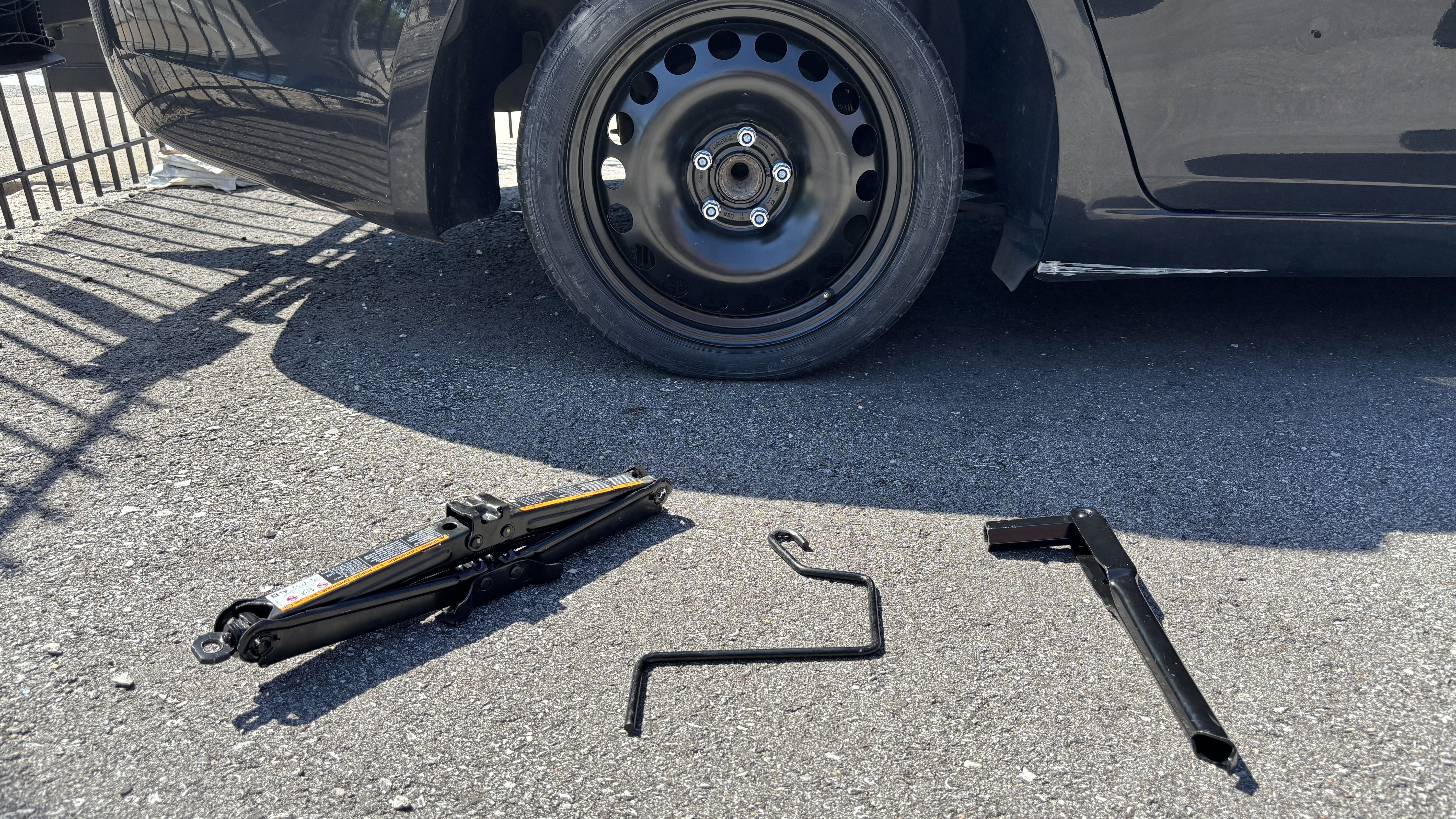 Black car with its front wheel removed, exposing a steel wheel and lug nuts. A hydraulic floor jack and tools lie on the asphalt, with fence shadows crossing the ground.