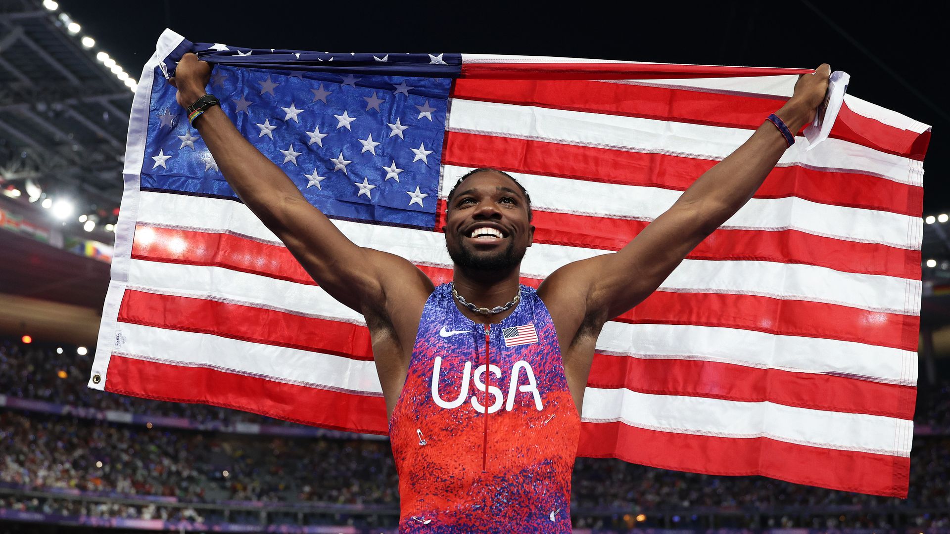 A man holds up the American flag behind him with both hands while smiling in a USA top with a crowd behind him.