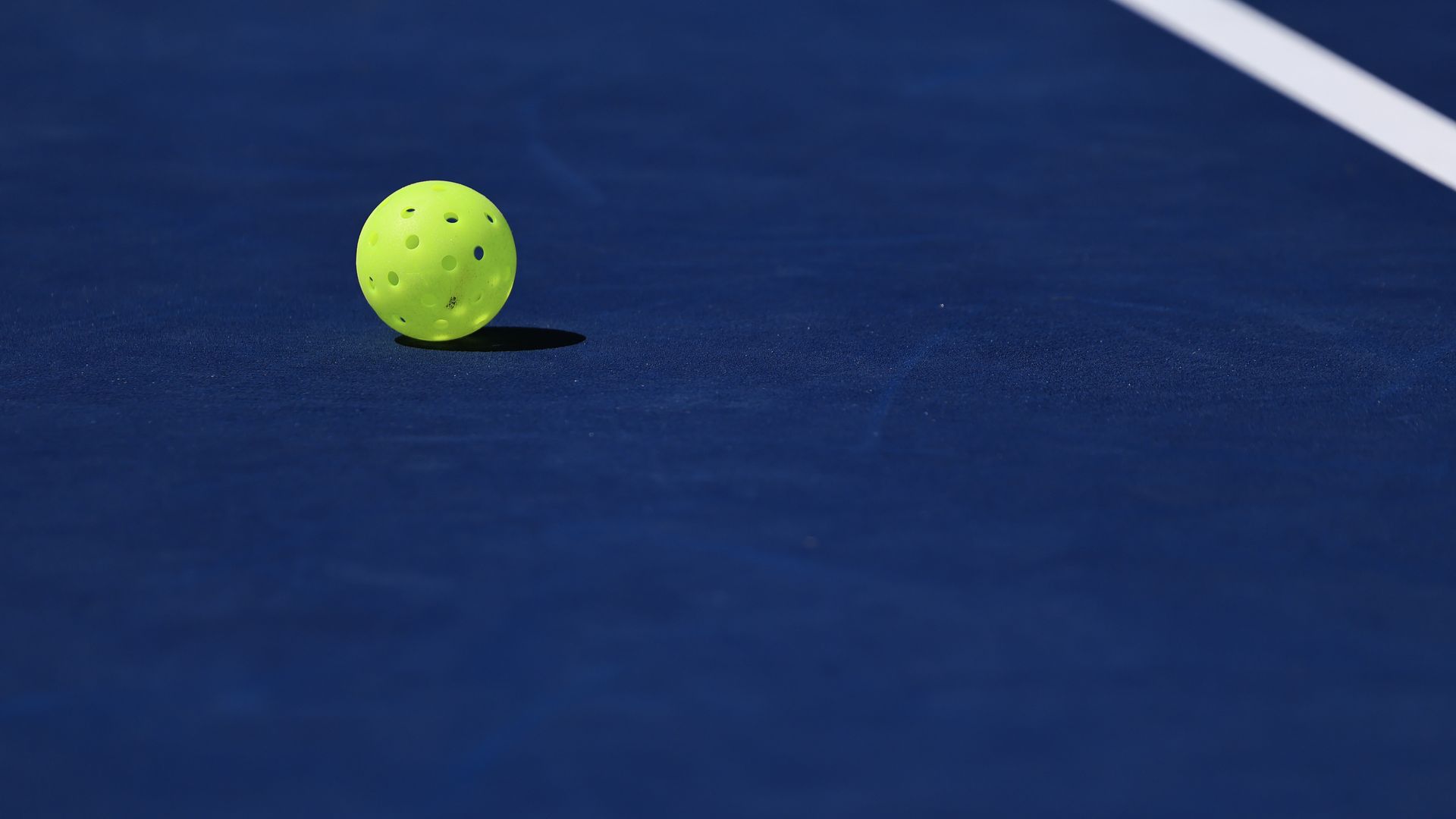 Neon green perforated ball rests on a dark blue court beside a white boundary line, casting a small shadow on the surface.