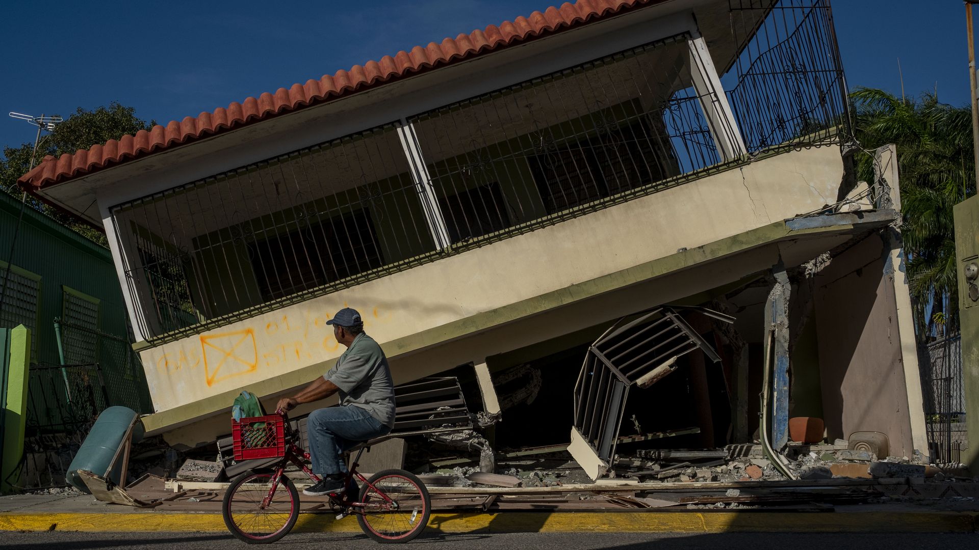 In this image, a Puerto Rican man bikes past a lopsided building. 