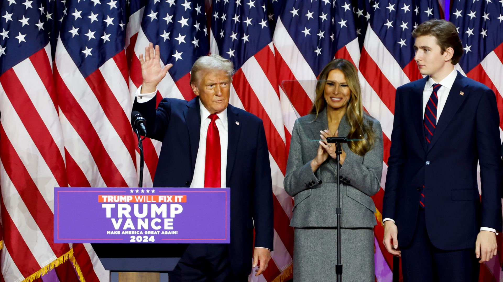 Donald Trump, Melania Trump and Barron Trump all stand in front of several American flags. 
