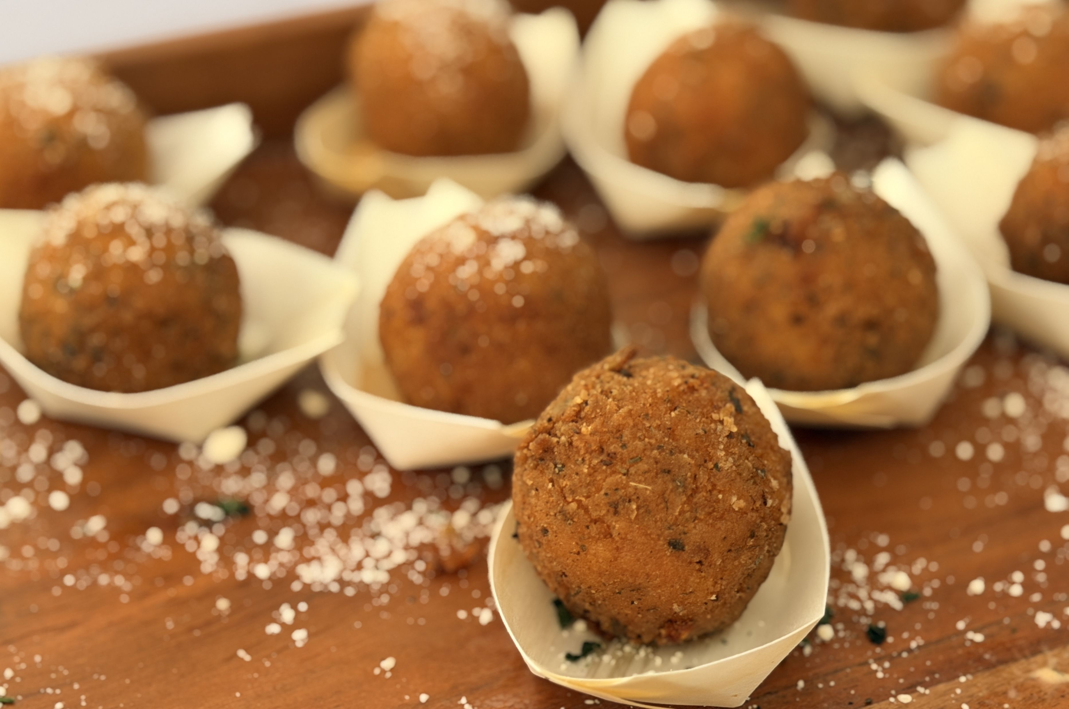 Several golden-brown, breaded croquettes sit in white paper boats on a wooden table, sprinkled with sesame seeds, with a glossy sauce in the background.