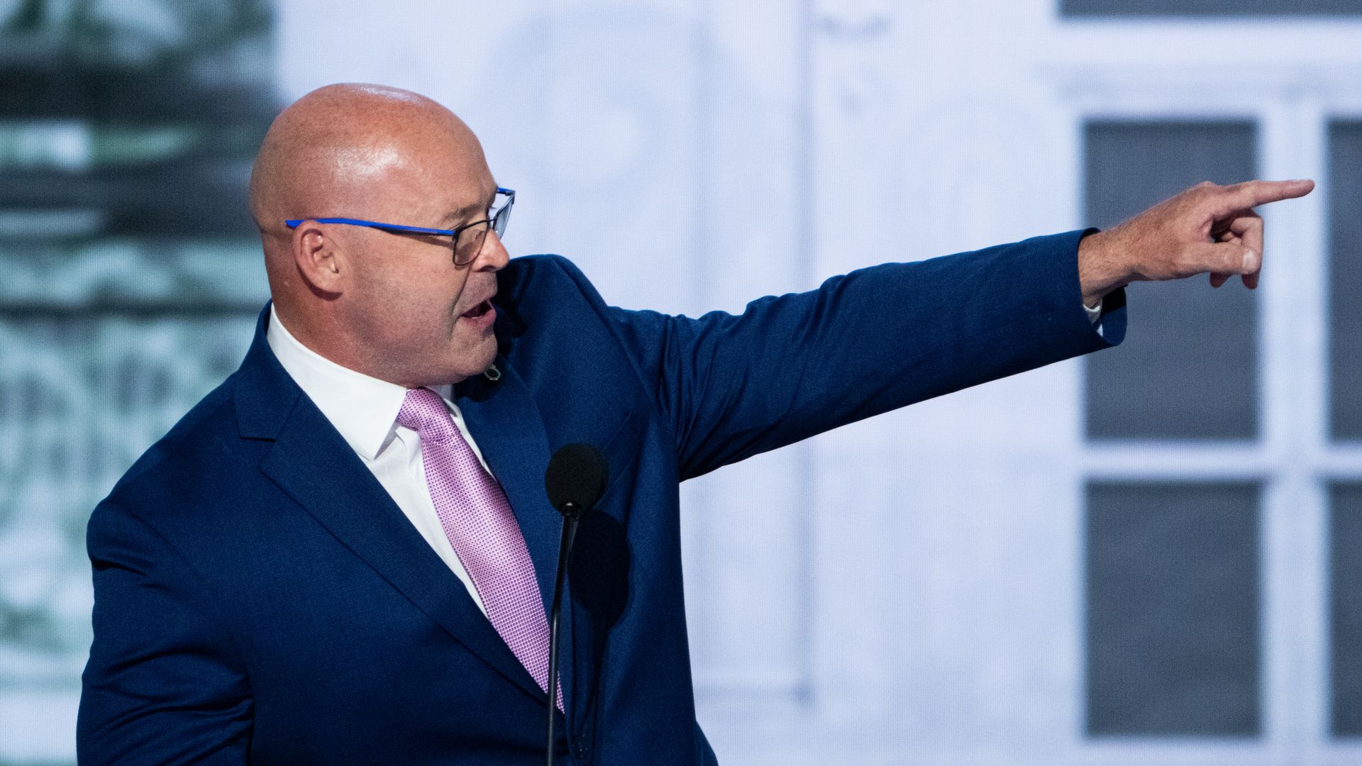A person in a dark blue suit, white collared shirt and pink tie with purple eyeglasses points with their left hand while speaking