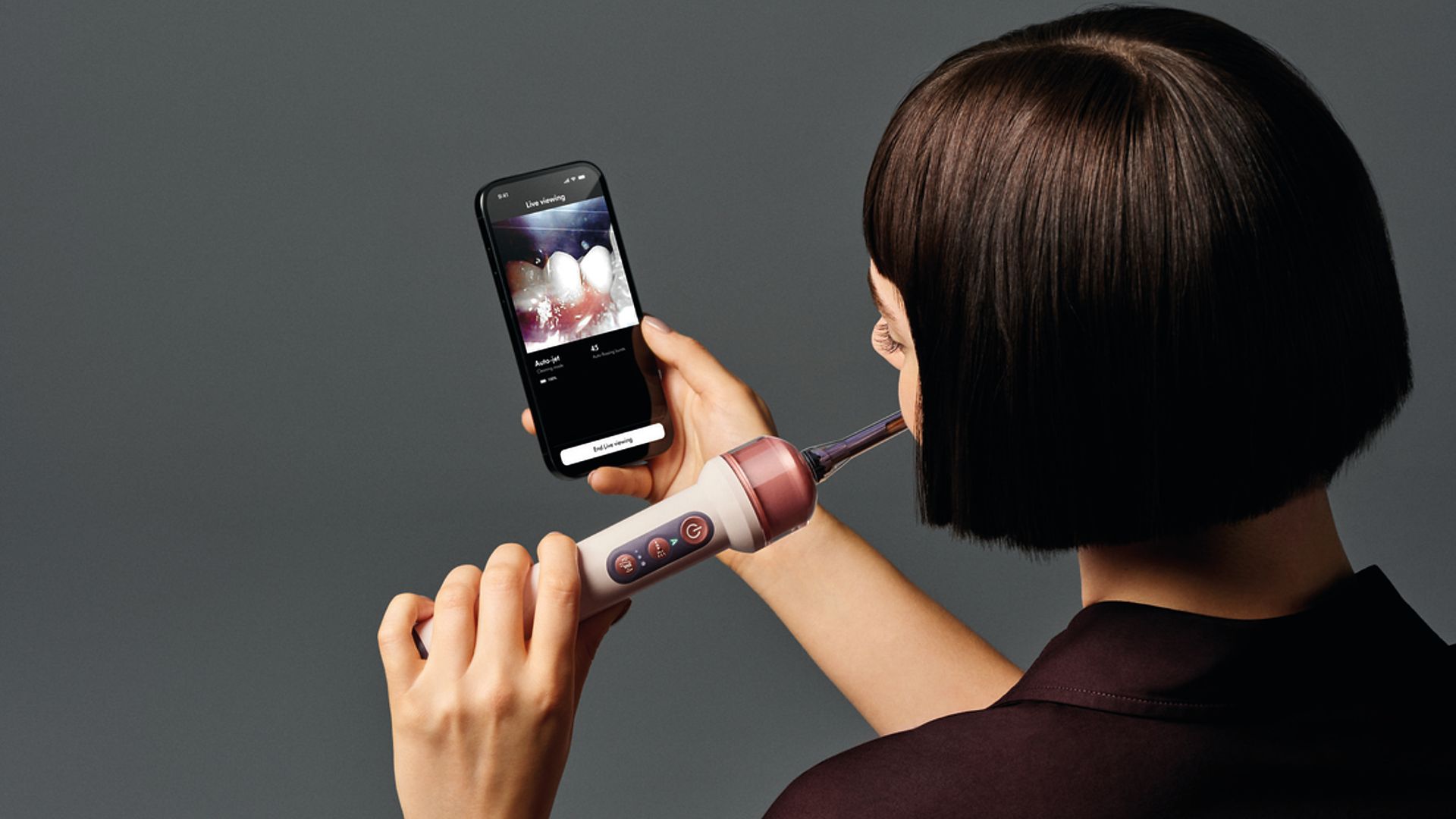 Person with a short dark bob uses a dental wand while viewing their mouth on a smartphone displaying a "live viewing" screen, against a gray background.