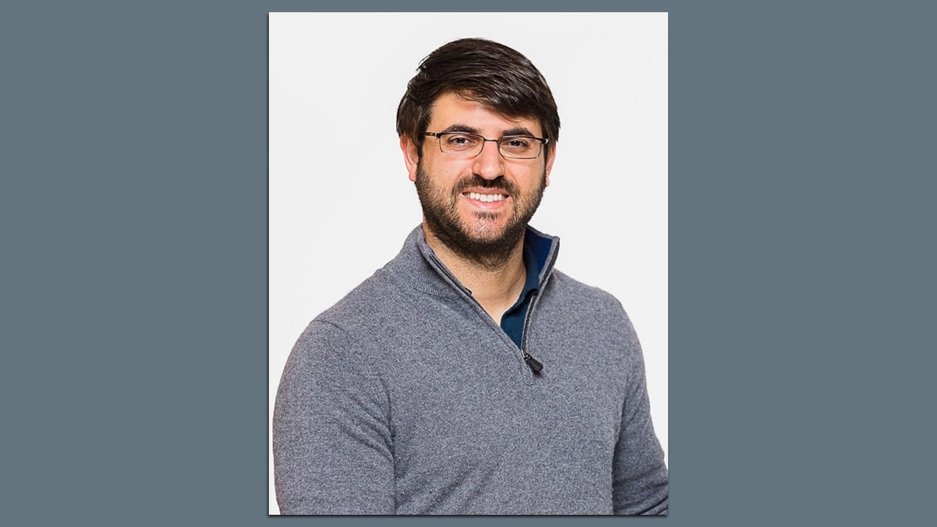 Smiling man with dark hair and glasses, wearing a gray quarter-zip sweater, posed against a plain light background.