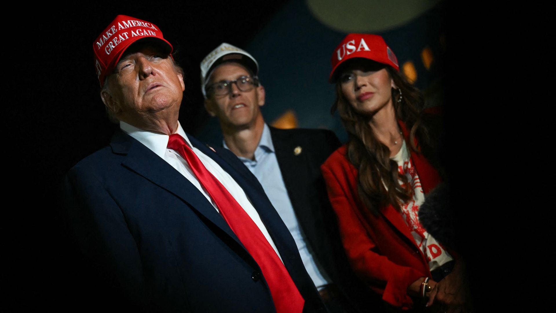 President Trump, wearing a red and white "MAGA" cap, navy suit, white shirt and red tie, a bespectacled man in a white and gold and cap, navy blazor and denim shirt, and Secretary of Homeland Security Kristi Noem, wearing a red and white "USA" cap, red blazor and yellow and red top.