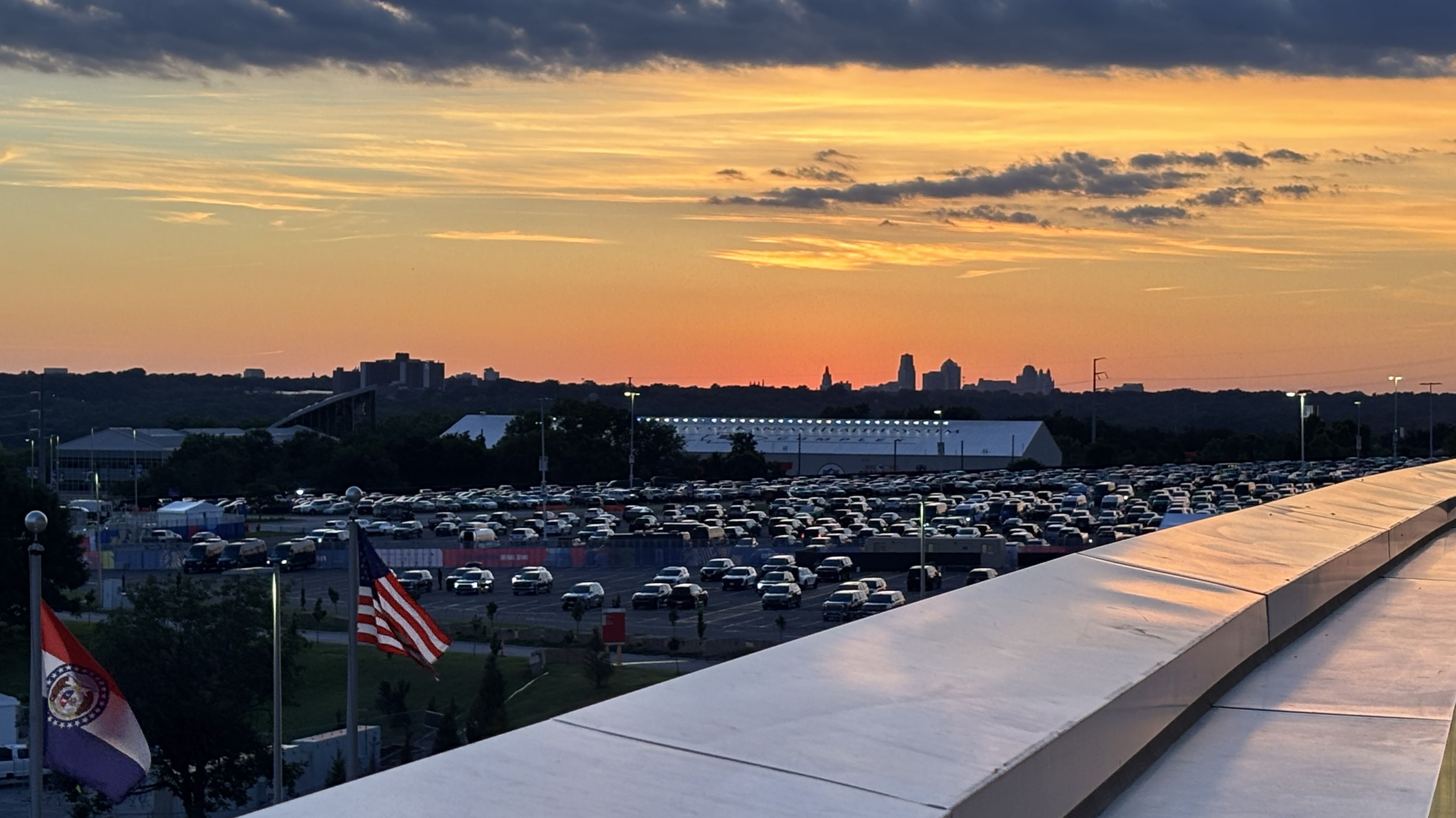 Sunset over a distant Kansas City skyline with orange and yellow clouds; in the foreground a large parking lot full of cars and a curved white rooftop, with American flags in the lower left.