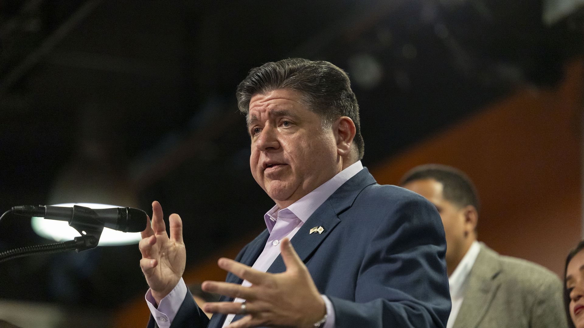 JB Pritzker, wearing a light shirt and a dark suit, gestures as he speaks into a microphone.