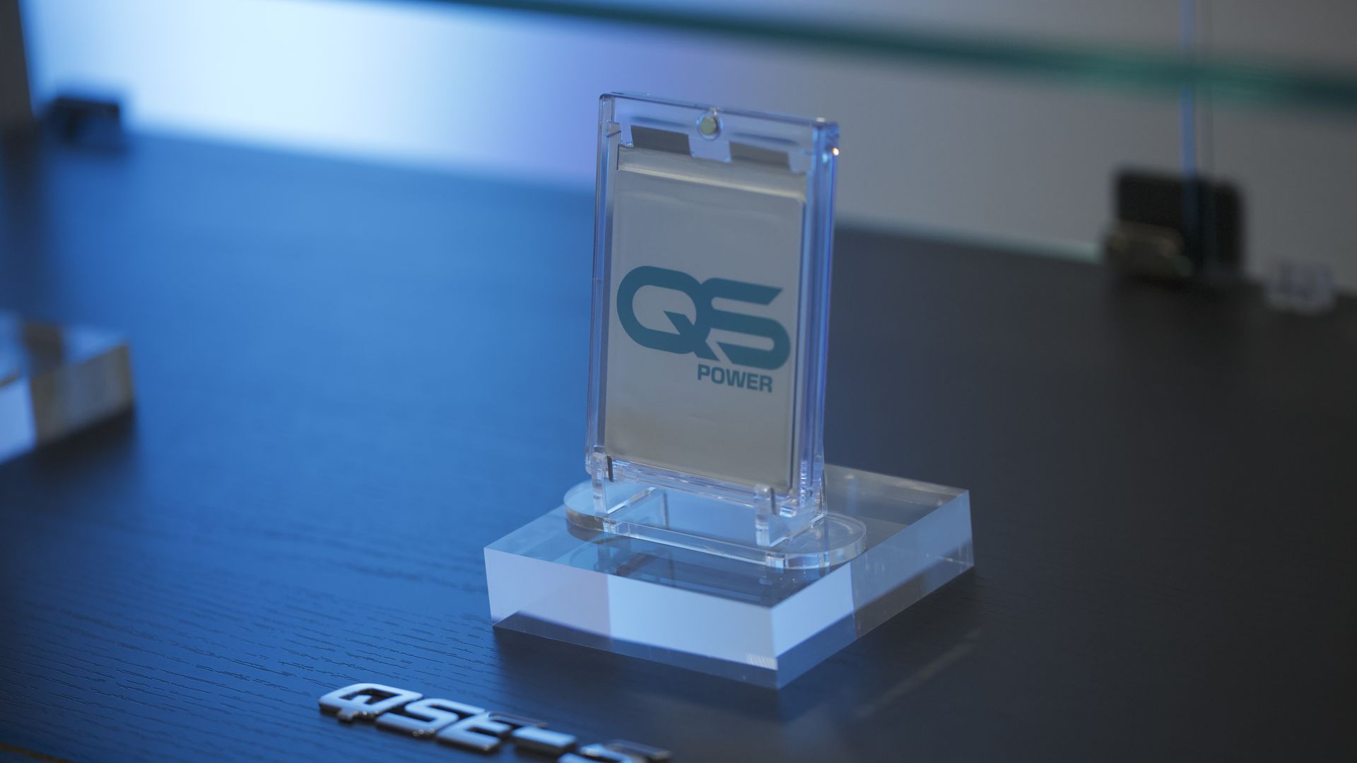 Clear acrylic display stand with a card inside showing the logo 'QS POWER' in teal on a dark wooden surface with a metallic QS logo nearby, lit with blue light.