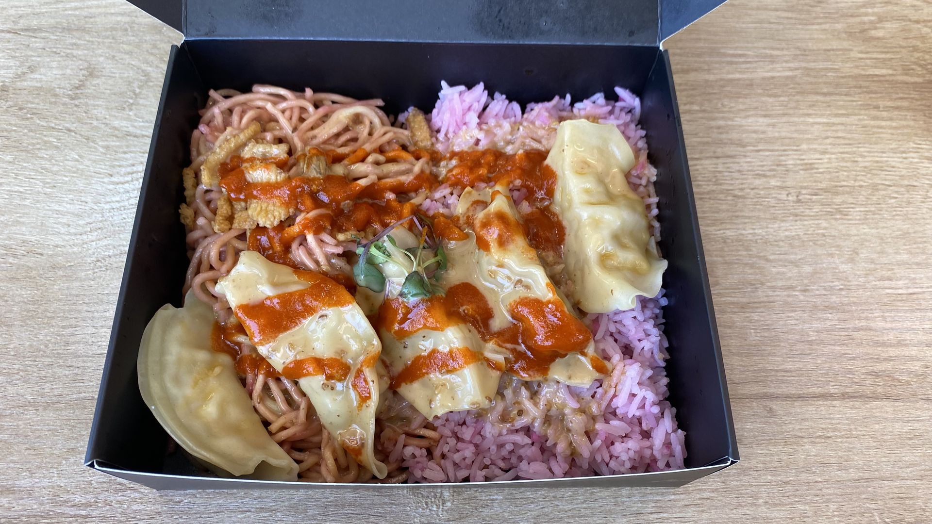 Open black takeout box on a wooden table, with noodles and pink rice, topped with dumplings with red sauced drizzled on them.