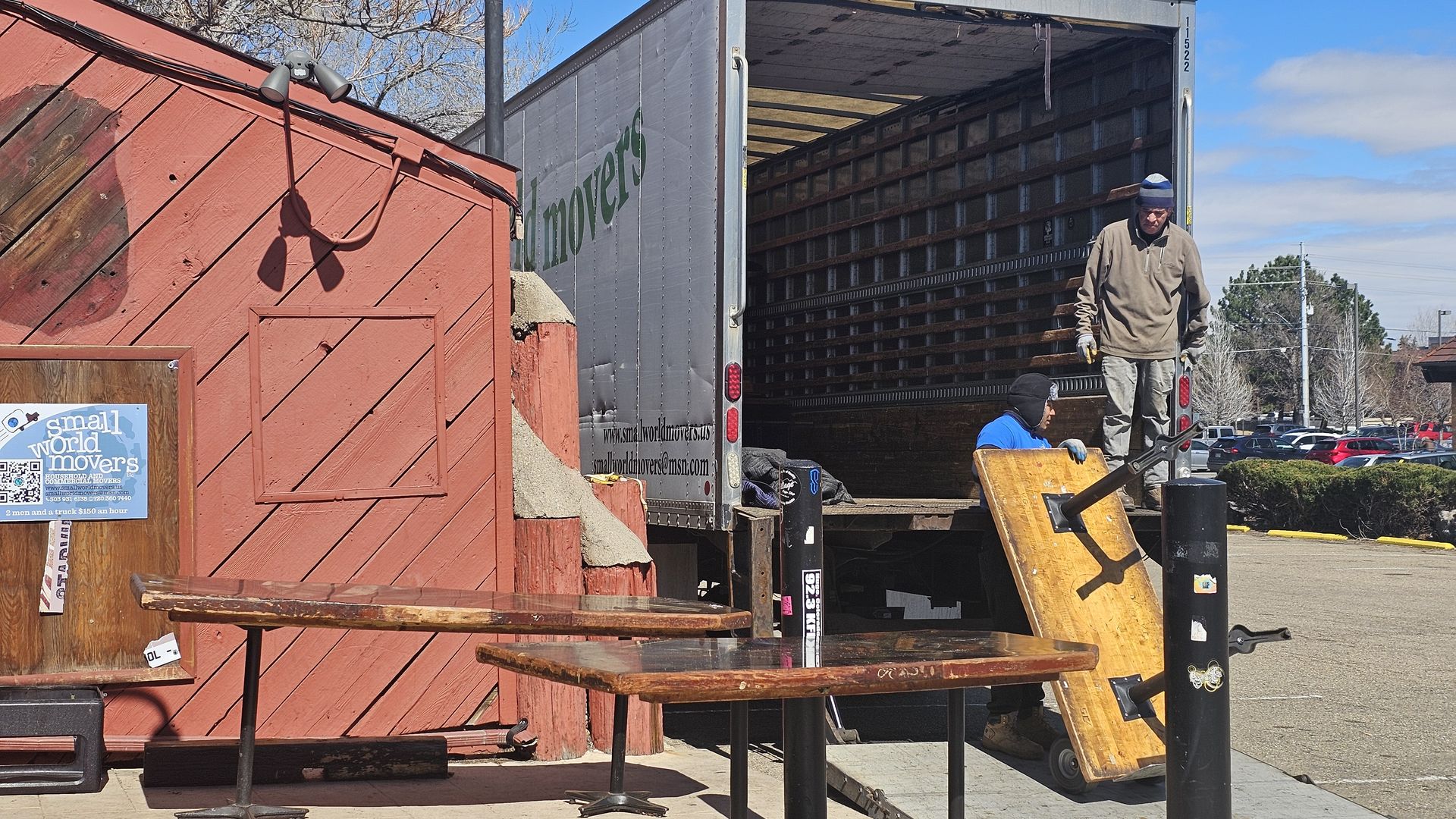 Two movers unload from an open-back moving truck in a parking lot. One stands on the ramp in a beige hoodie, another in blue on the ground near a red wooden building with a Small World Movers sign.