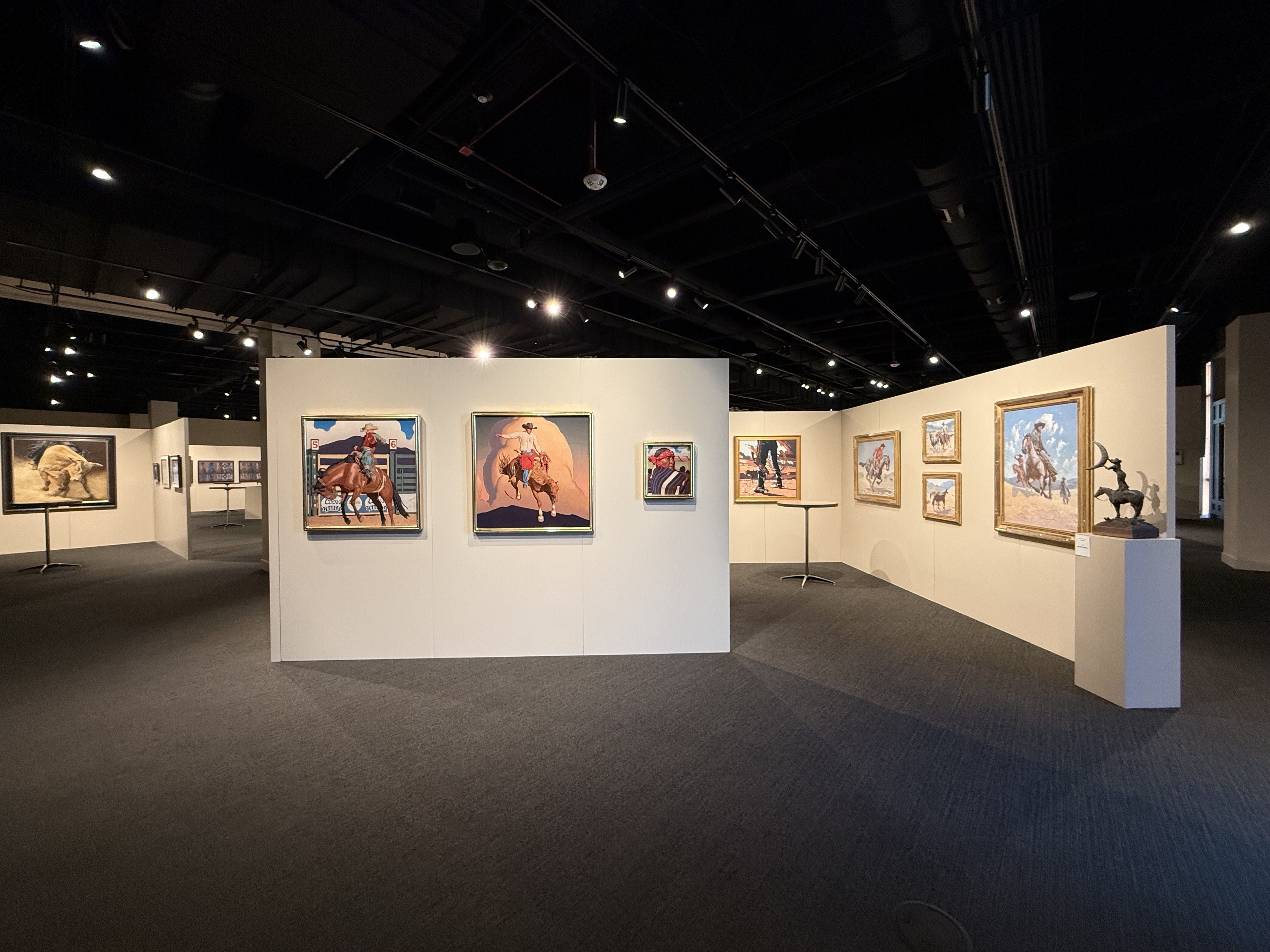 Art gallery interior featuring paintings of cowboys and horses with a bronze cowboy sculpture on a pedestal under soft lighting.