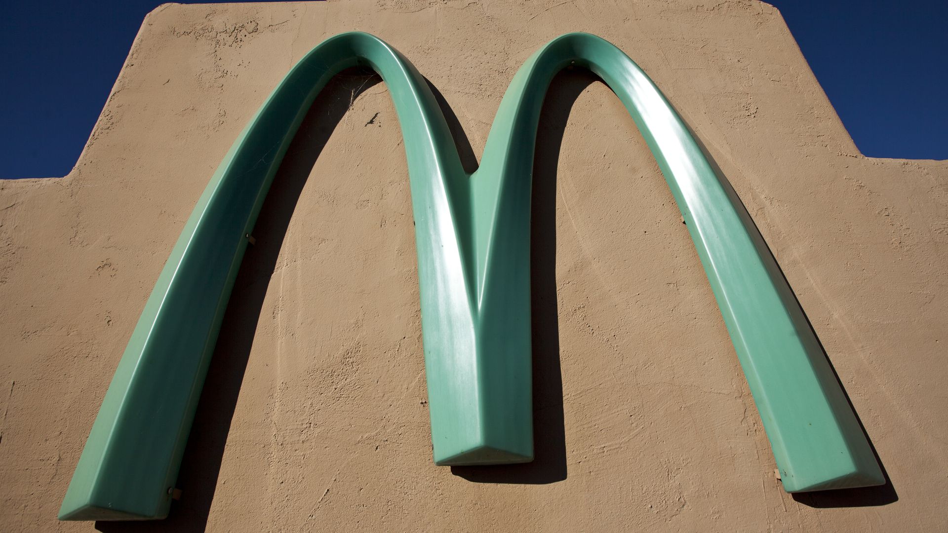 Close-up of two turquoise-green curved metal arches forming an M against a beige stucco wall under a clear blue sky.
