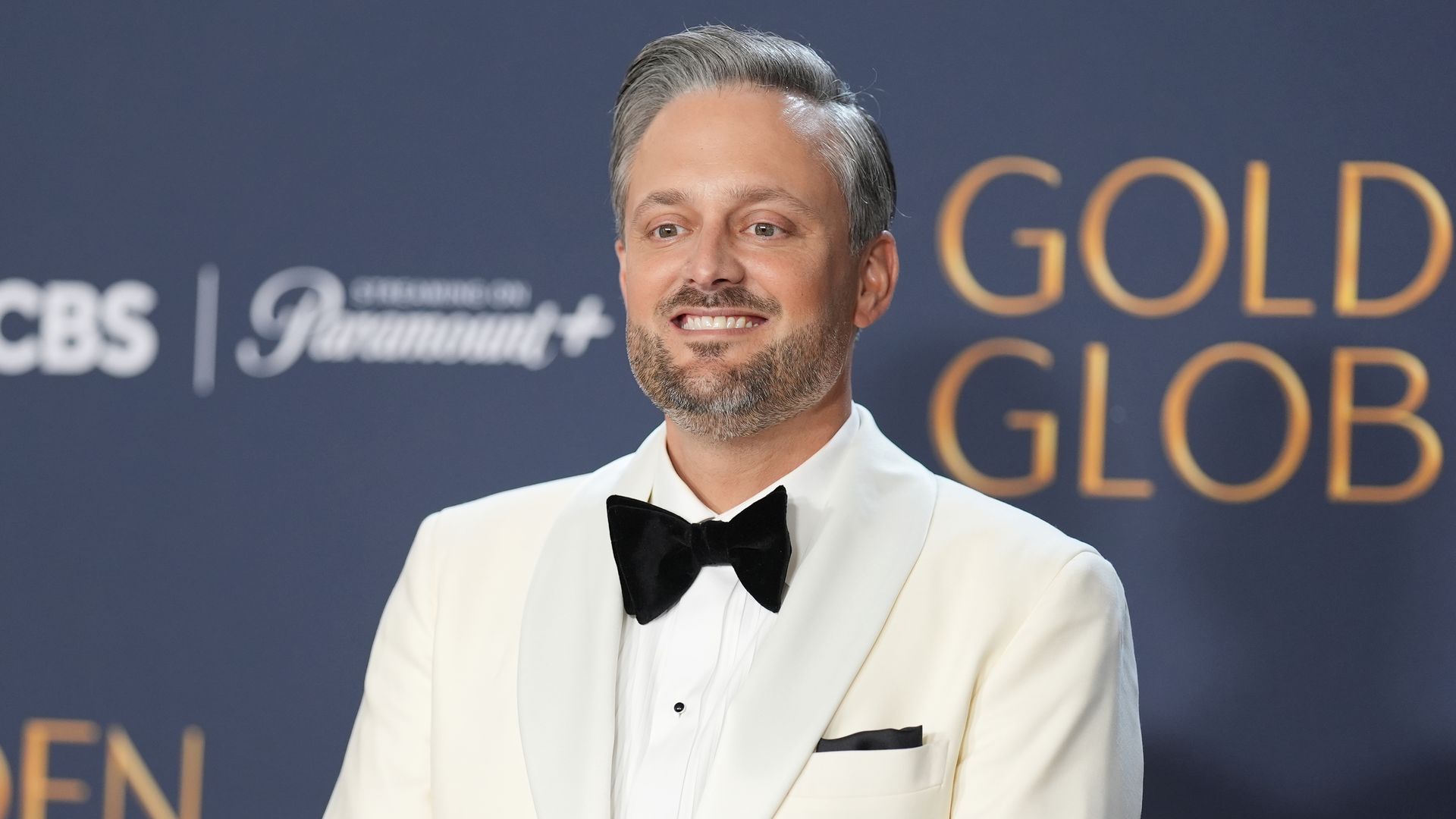 A man wearing a white tuxedo jacket with a black bow tie and pocket square poses on the red carpet at the Golden Globe Awards, smiling in front of a backdrop with the event’s logo.