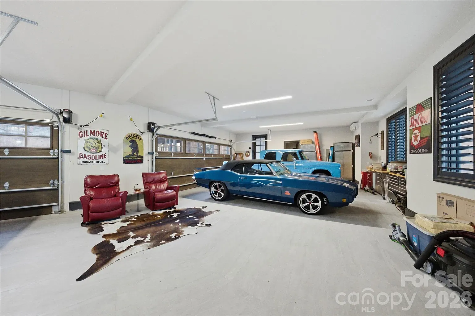Bright garage with a blue classic convertible center, a light-blue truck in back, two red leather chairs on a cowhide rug, vintage gas signs on the walls, and a workbench to the right.