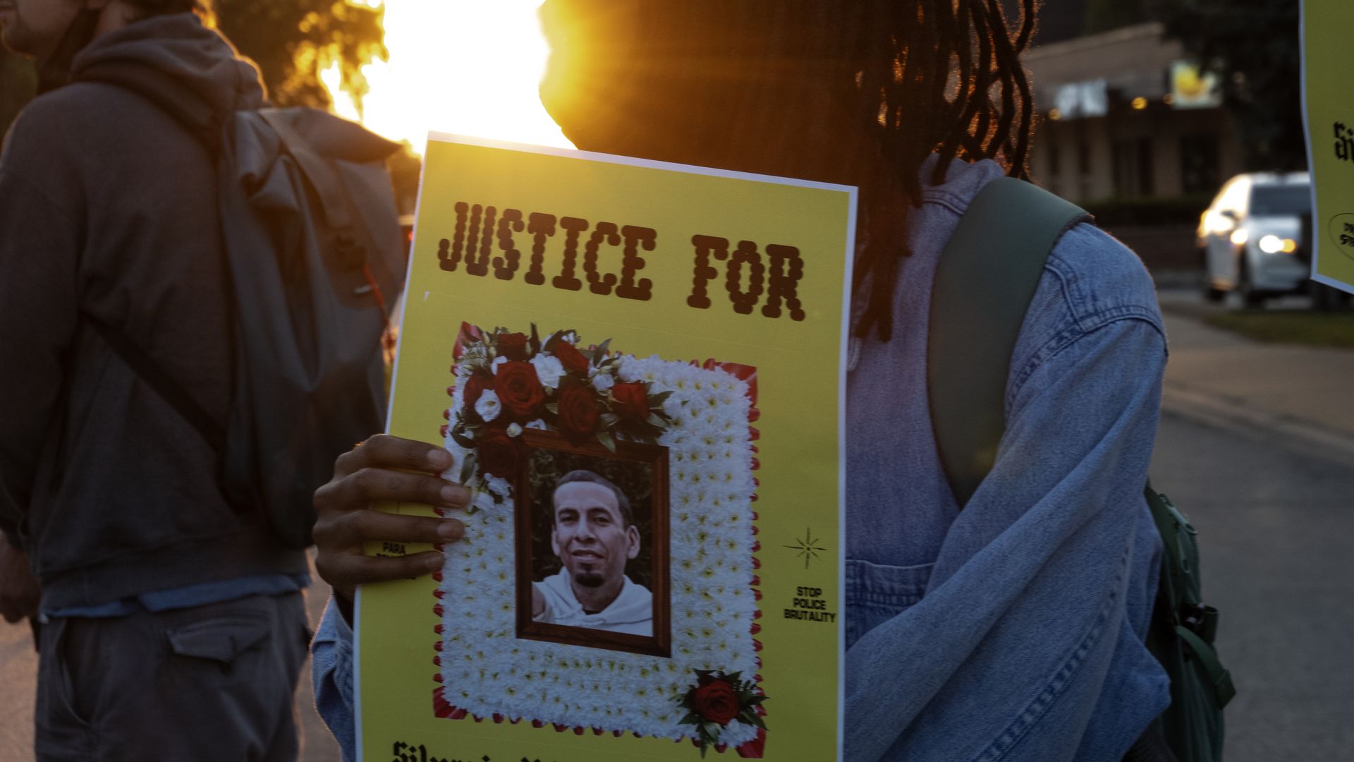Back of a protester with dreadlocks in a denim jacket, holding a bright yellow poster that reads "JUSTICE FOR" with a flower wreath and portrait; sunset glow on a city street.