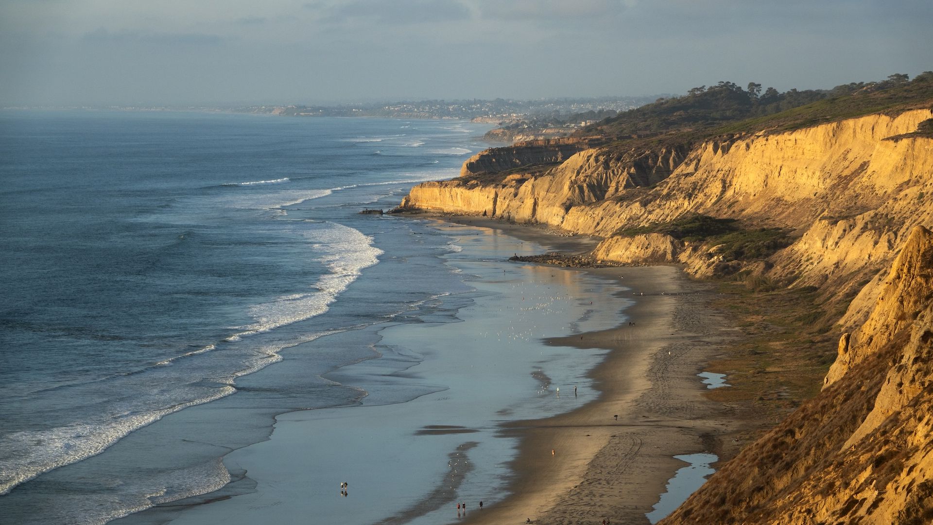 The sun shines on the bluffs of Black's Beach and Torrey Pines State Natural Reserve as waves in the Pacific Ocean roll in at low tide.