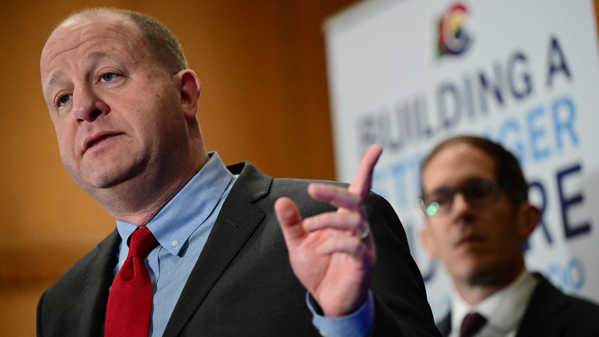 A man in a suit and necktie points outwardly while speaking. 