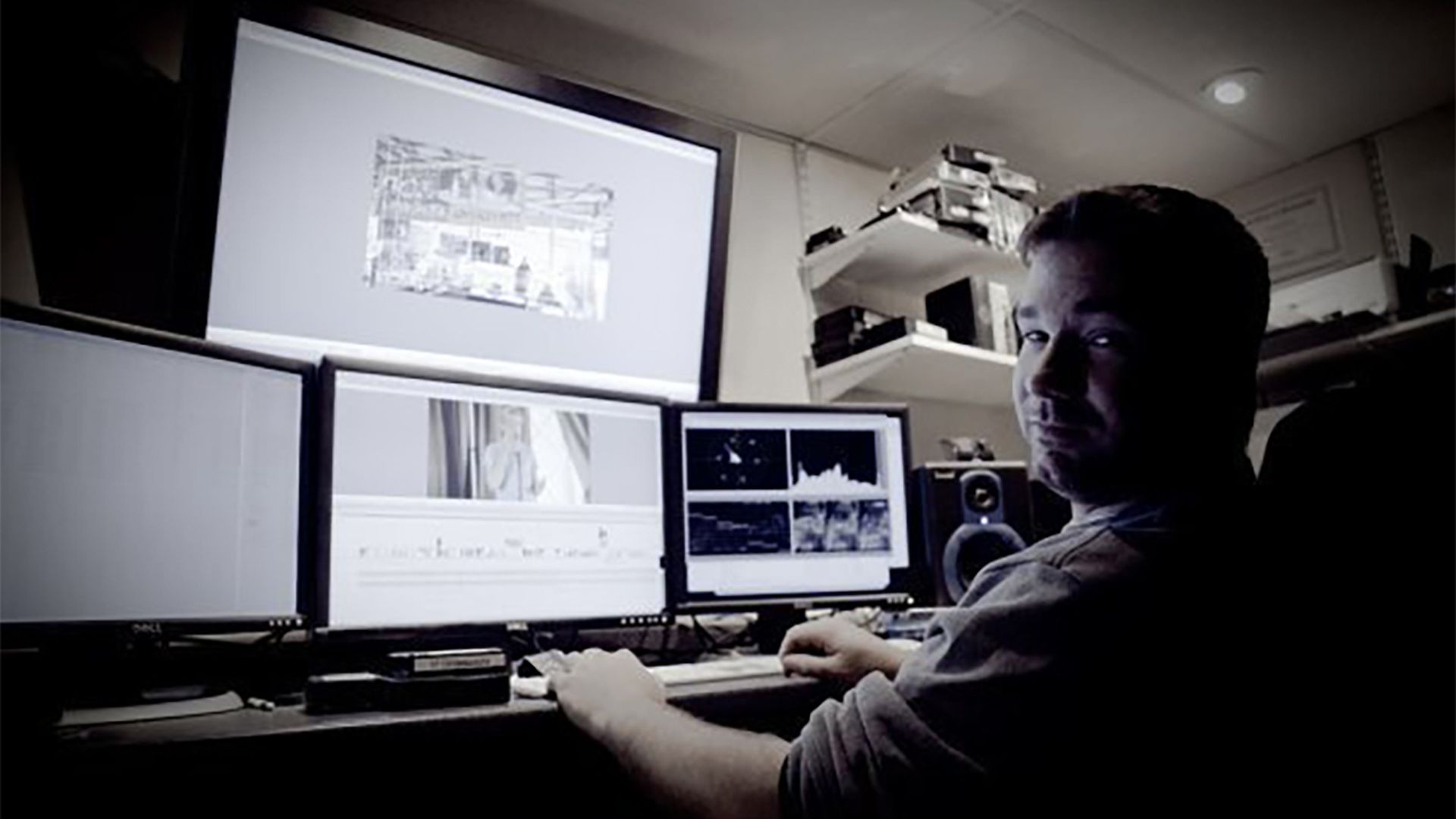 Man sitting at a desk with four computer monitors displaying video editing software and graphs, in a dimly lit room with shelves holding books and audio equipment.