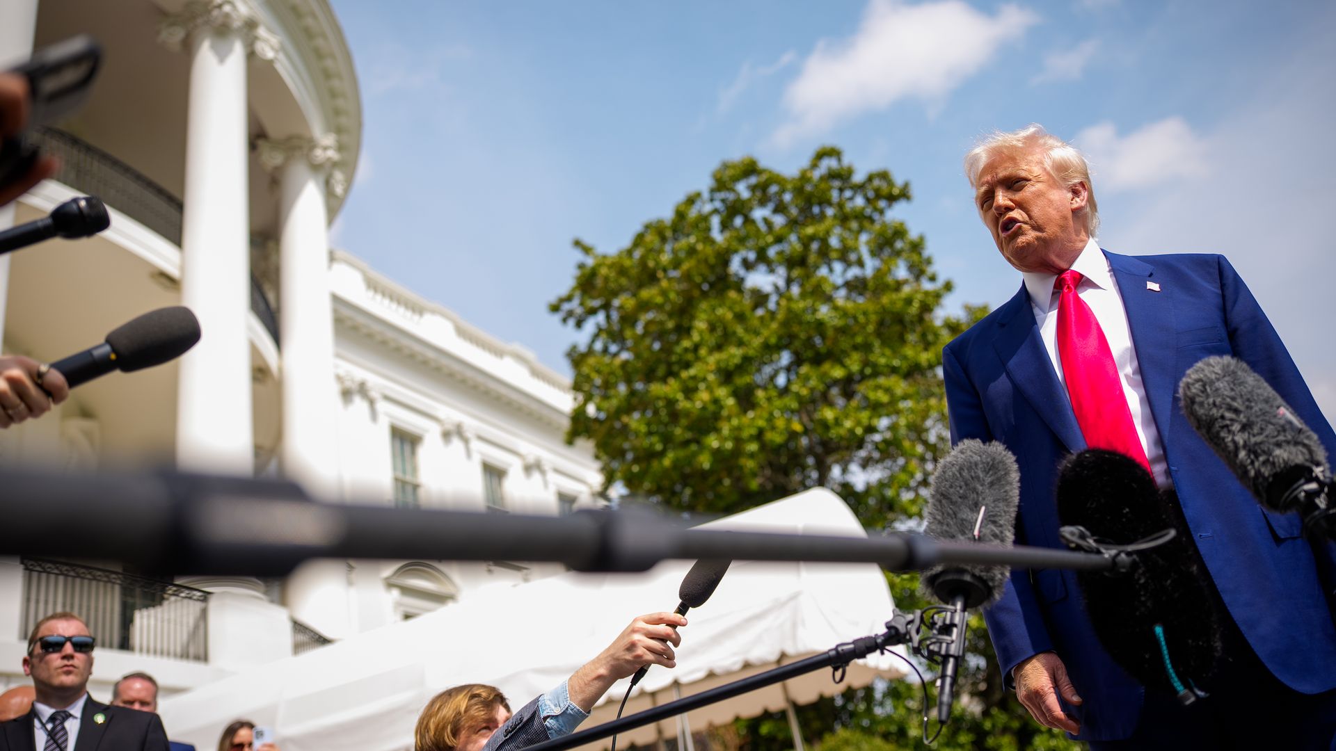 Trump, wearing a blue suit, white shirt and red tie, answers questions into several microphones while standing outside 