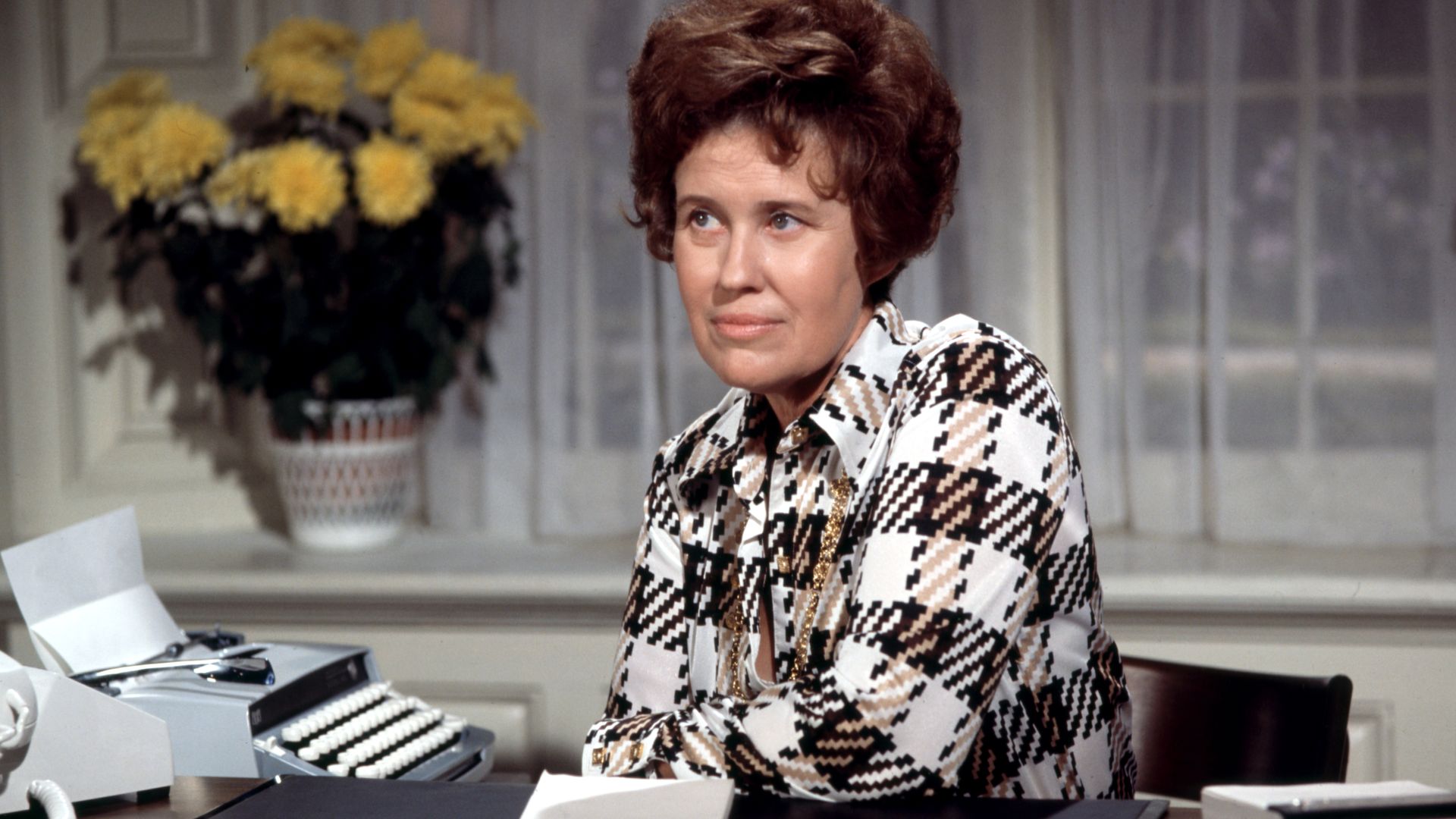 Erma Bombeck sitting at a table next to a typewriter and telephone. 